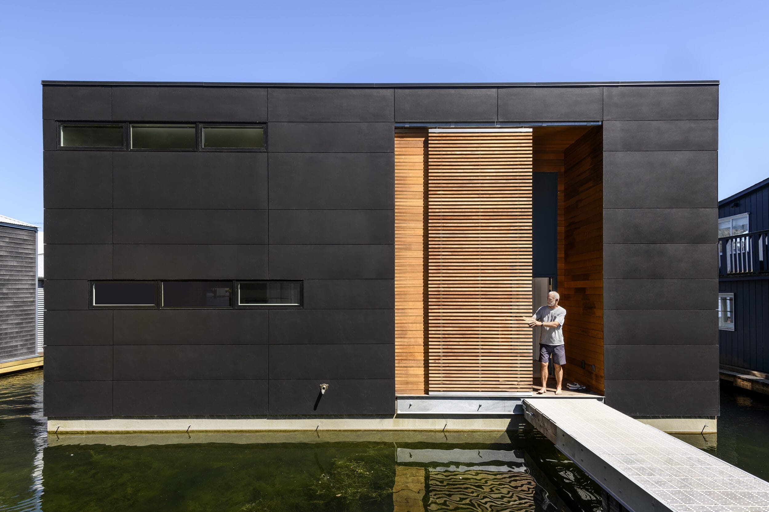 Modern black house on water with wooden accents, featuring a man standing at the entrance.