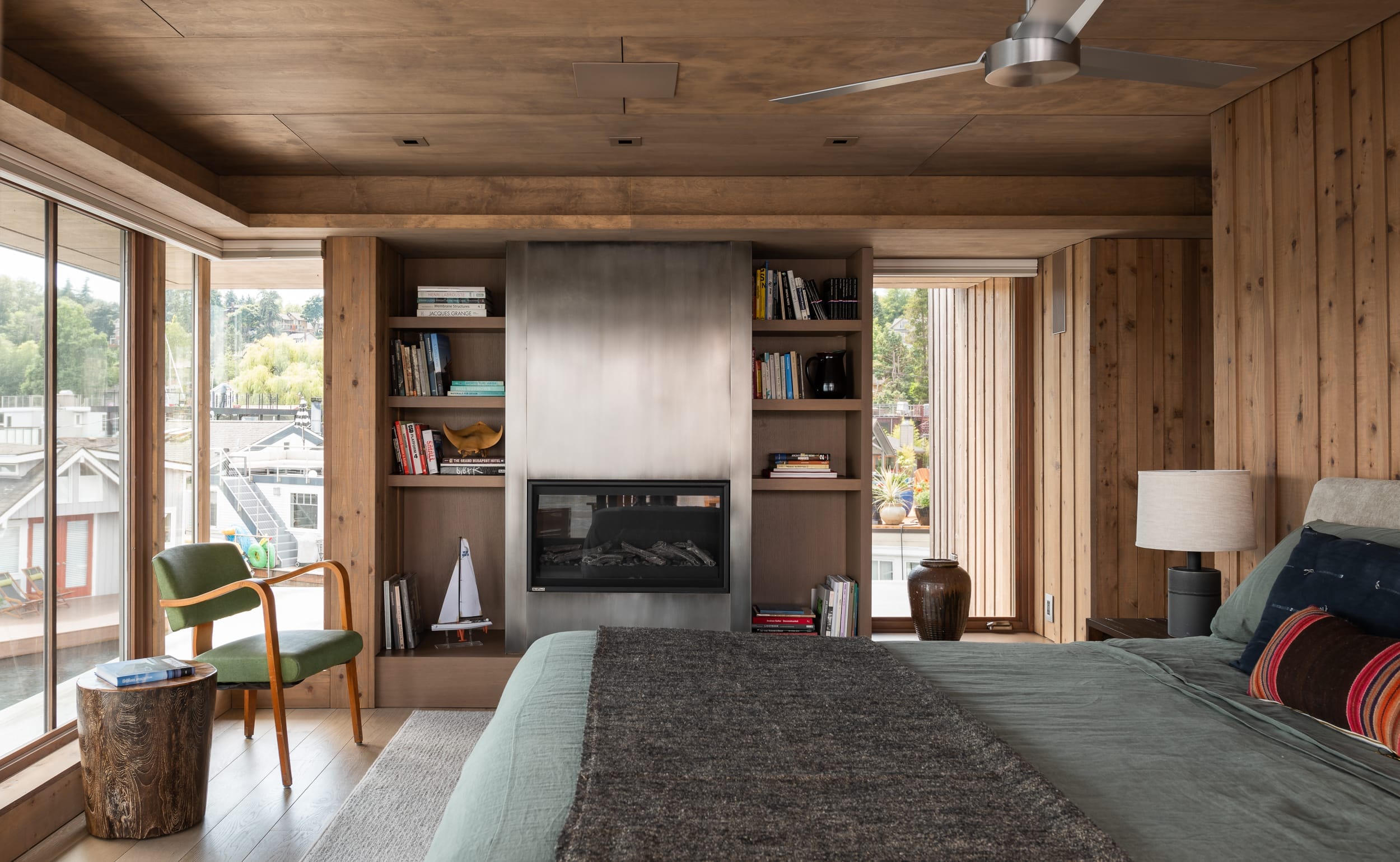 Cozy bedroom with wooden walls, a bed, an armchair, and a fireplace surrounded by shelves. Large windows offer a view of trees and buildings outside.