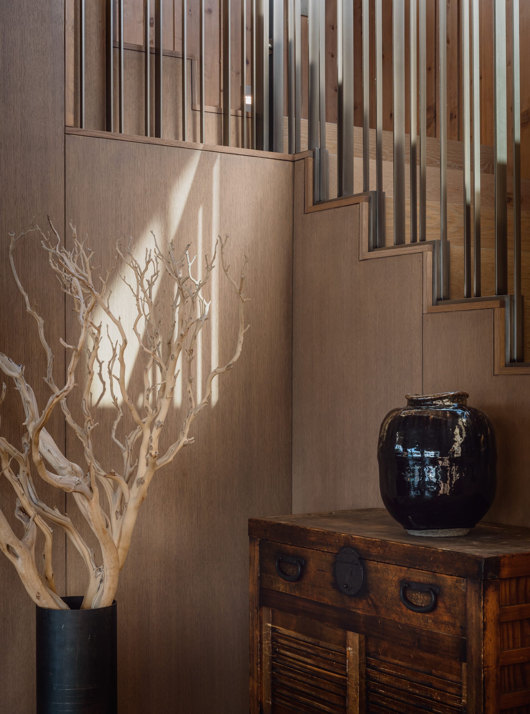 A wooden staircase with metal railings, a dark wooden cabinet, a large black vase, and a decorative dry branch in a pot are featured in the image.