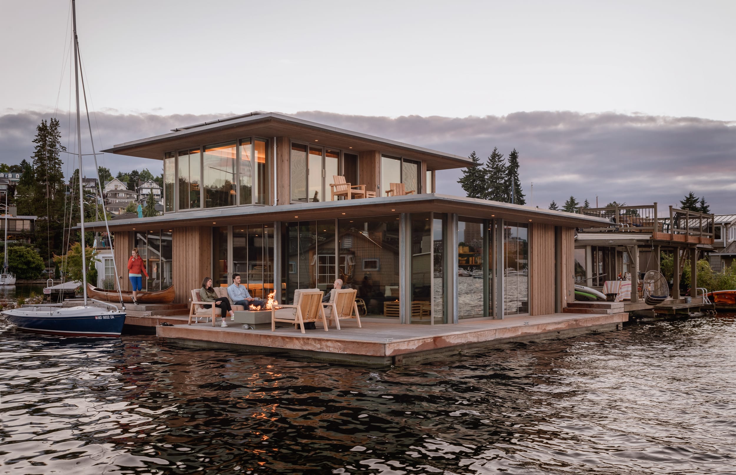 Modern floating house on the water, surrounded by trees and sailboats, with people sitting on the deck and enjoying an outdoor fire pit.