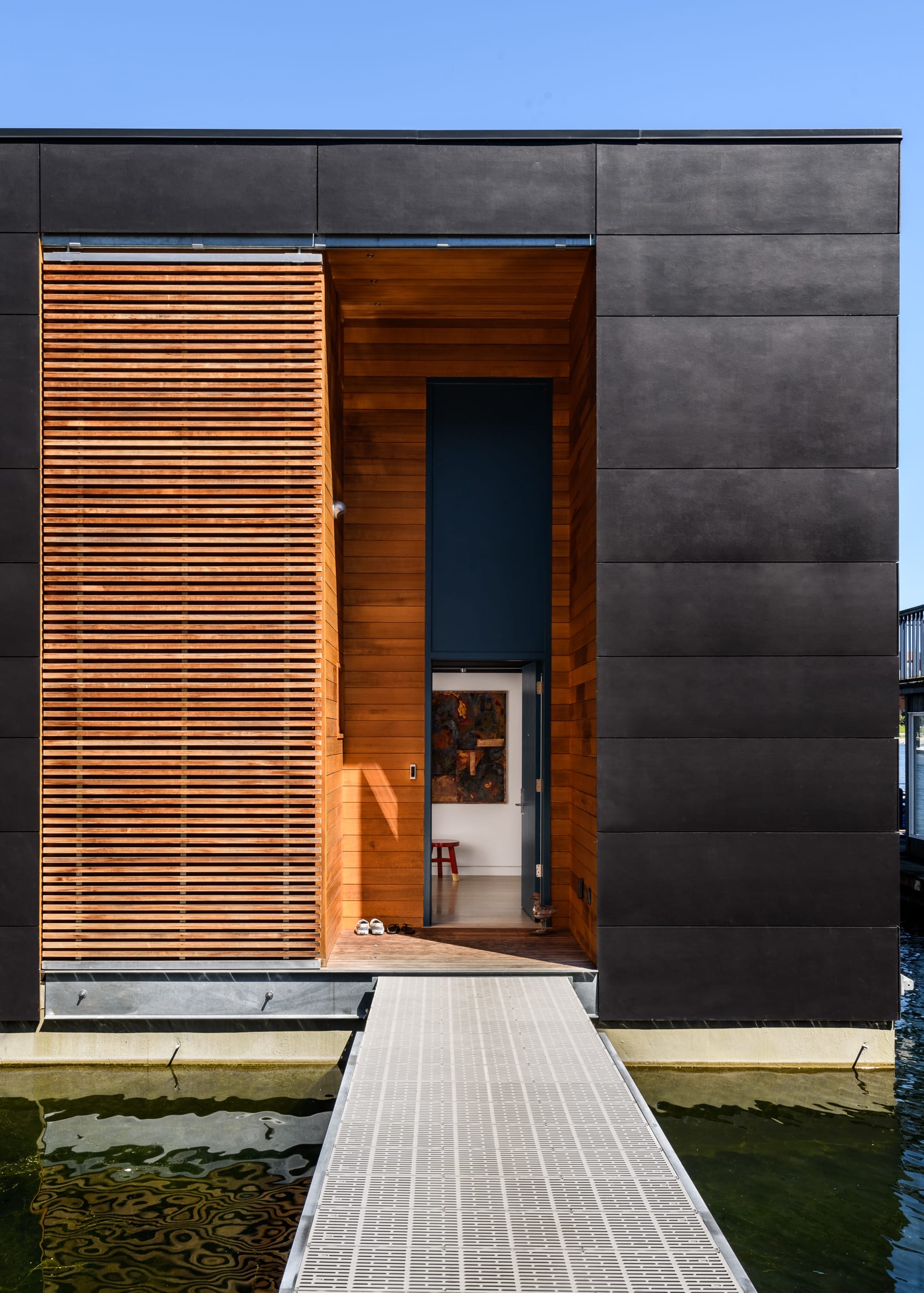 A modern floating house with a black facade, wooden slats, and a dock entrance extending over the water, viewed on a clear day.