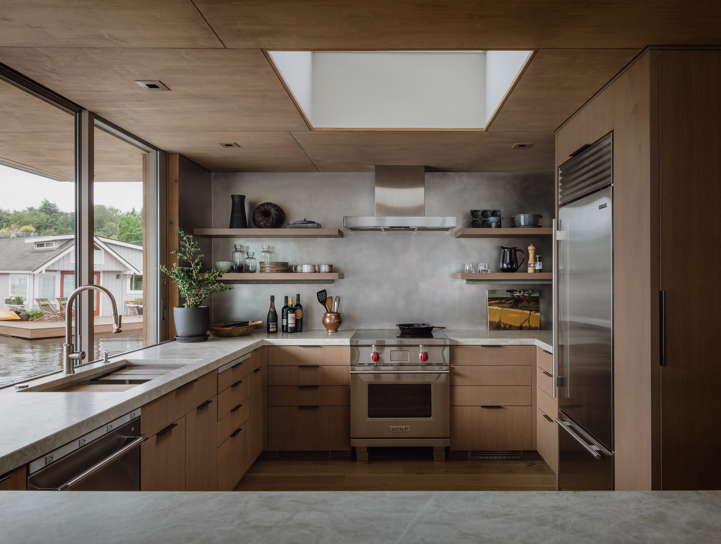 Modern kitchen with wooden cabinets, stainless steel appliances, open shelving, and a large window. A plant and various kitchen items are displayed on the shelves and counter.