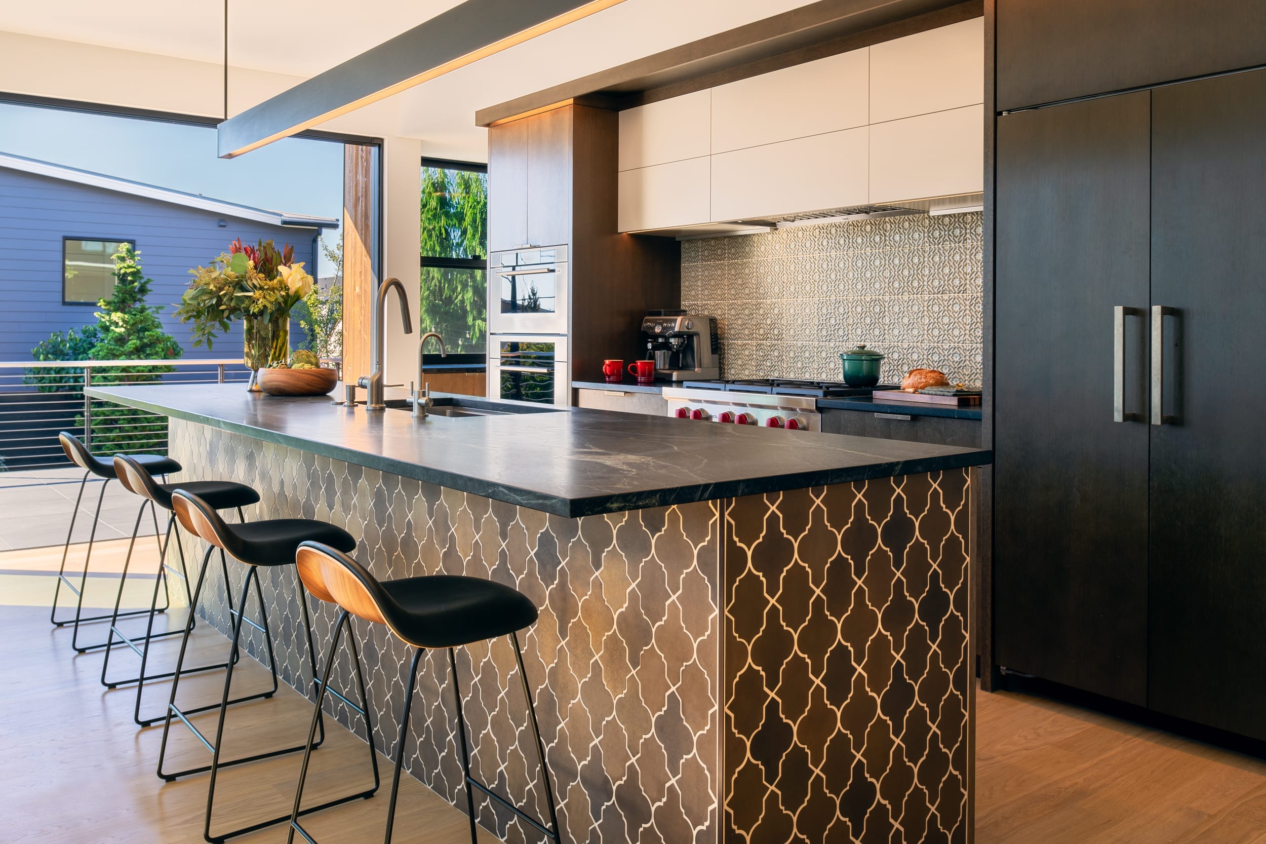 Modern kitchen with a geometric-patterned island, bar stools, and stainless steel appliances, featuring a large window overlooking greenery.