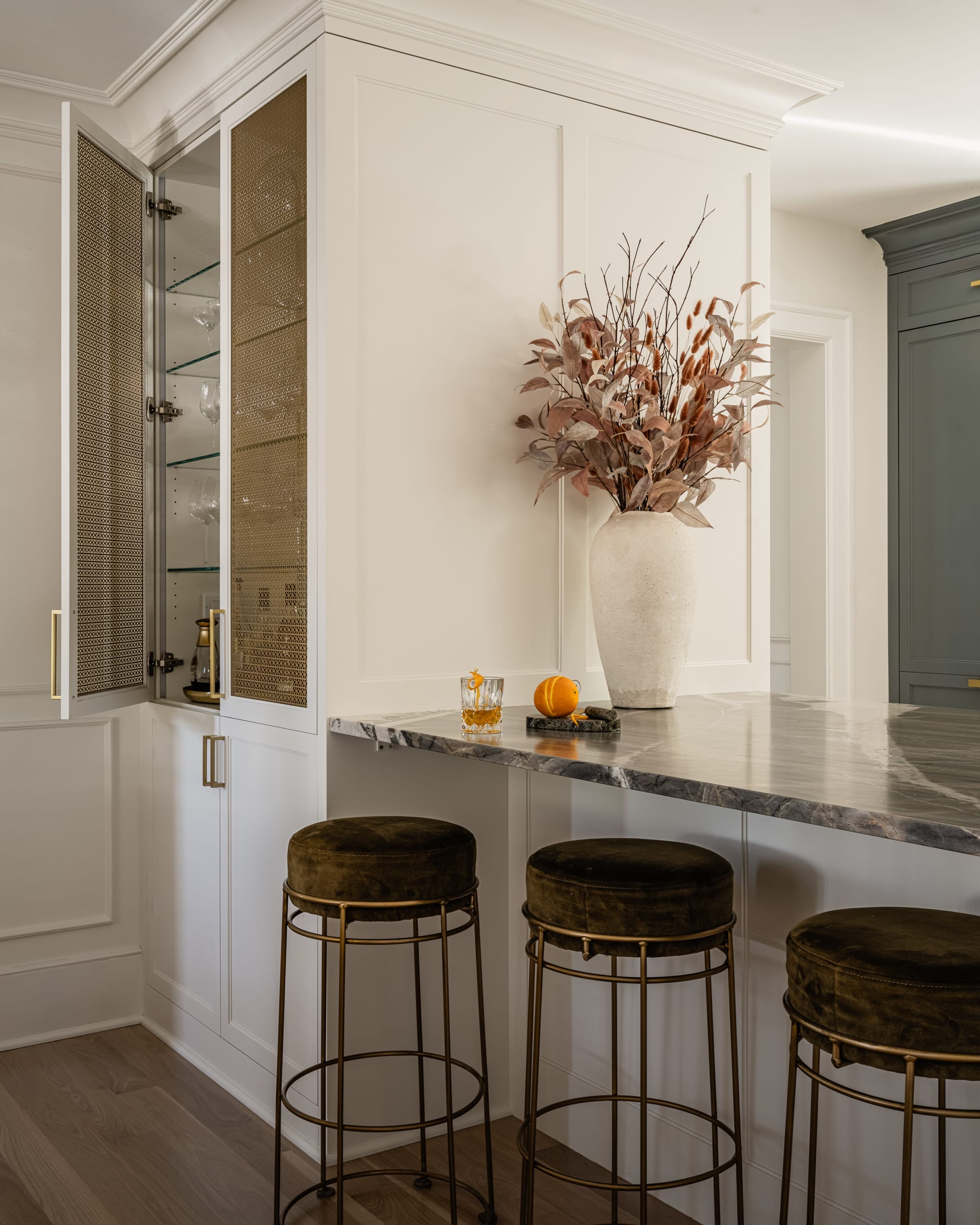 A kitchen with a marble countertop, bar stools, a large vase with dried leaves, a glass of whiskey, an orange, and an open cabinet displaying glassware.