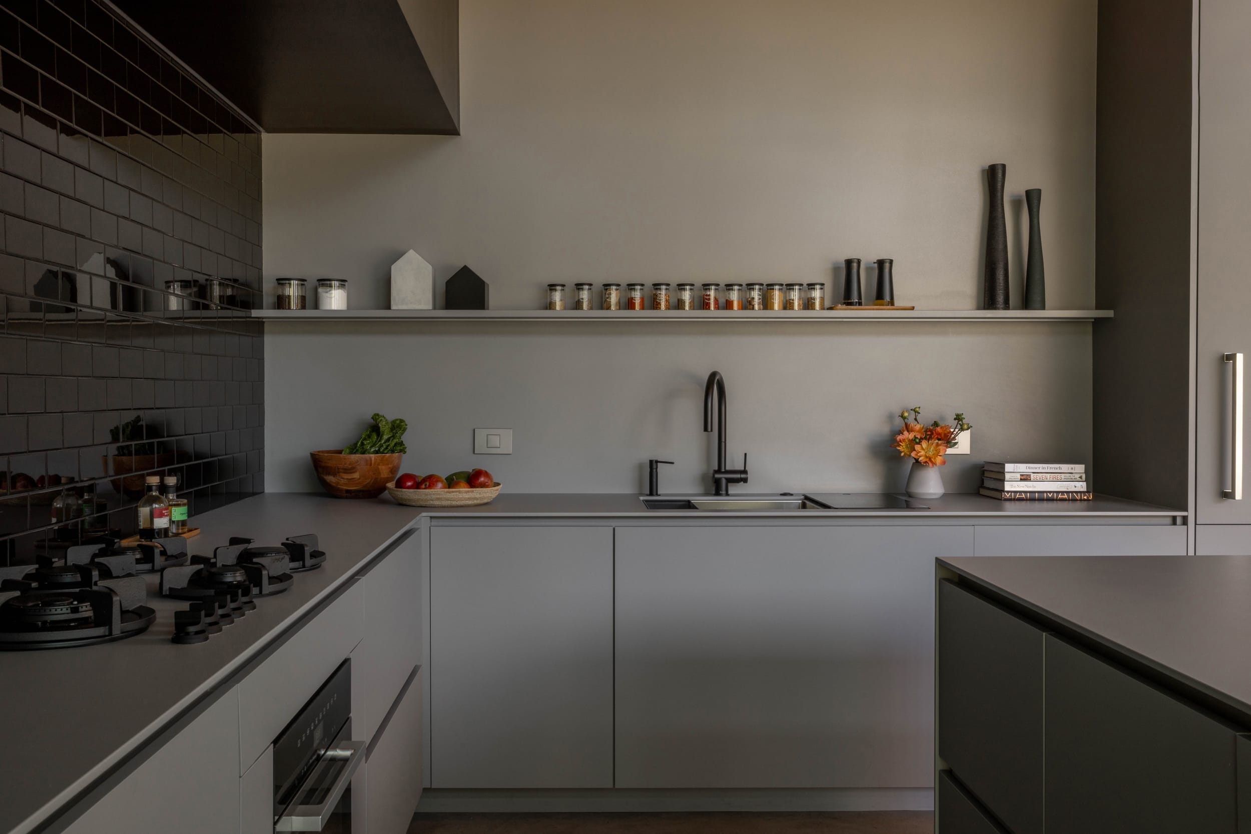 Modern kitchen with grey cabinets, black countertop, and black backsplash. Shelves hold spices and decor. Sink and stove are visible. Warm lighting adds a cozy ambiance.