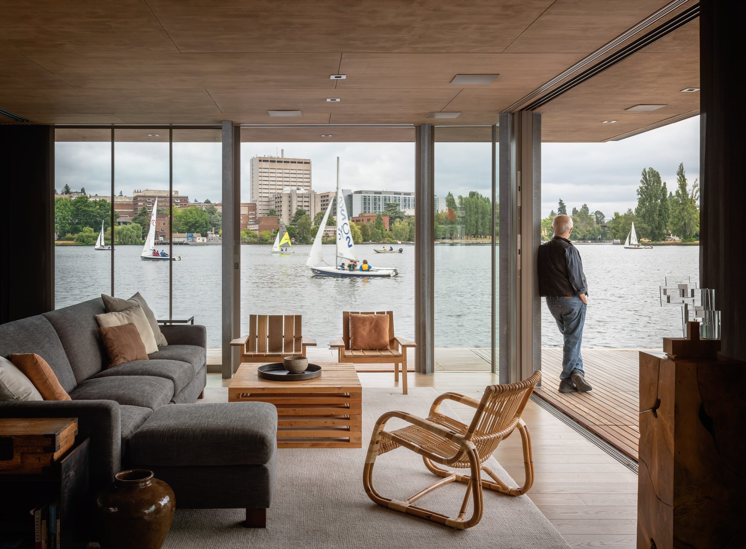 A man stands on a deck overlooking a lake with sailboats. The interior features modern furniture with large windows offering a scenic view.