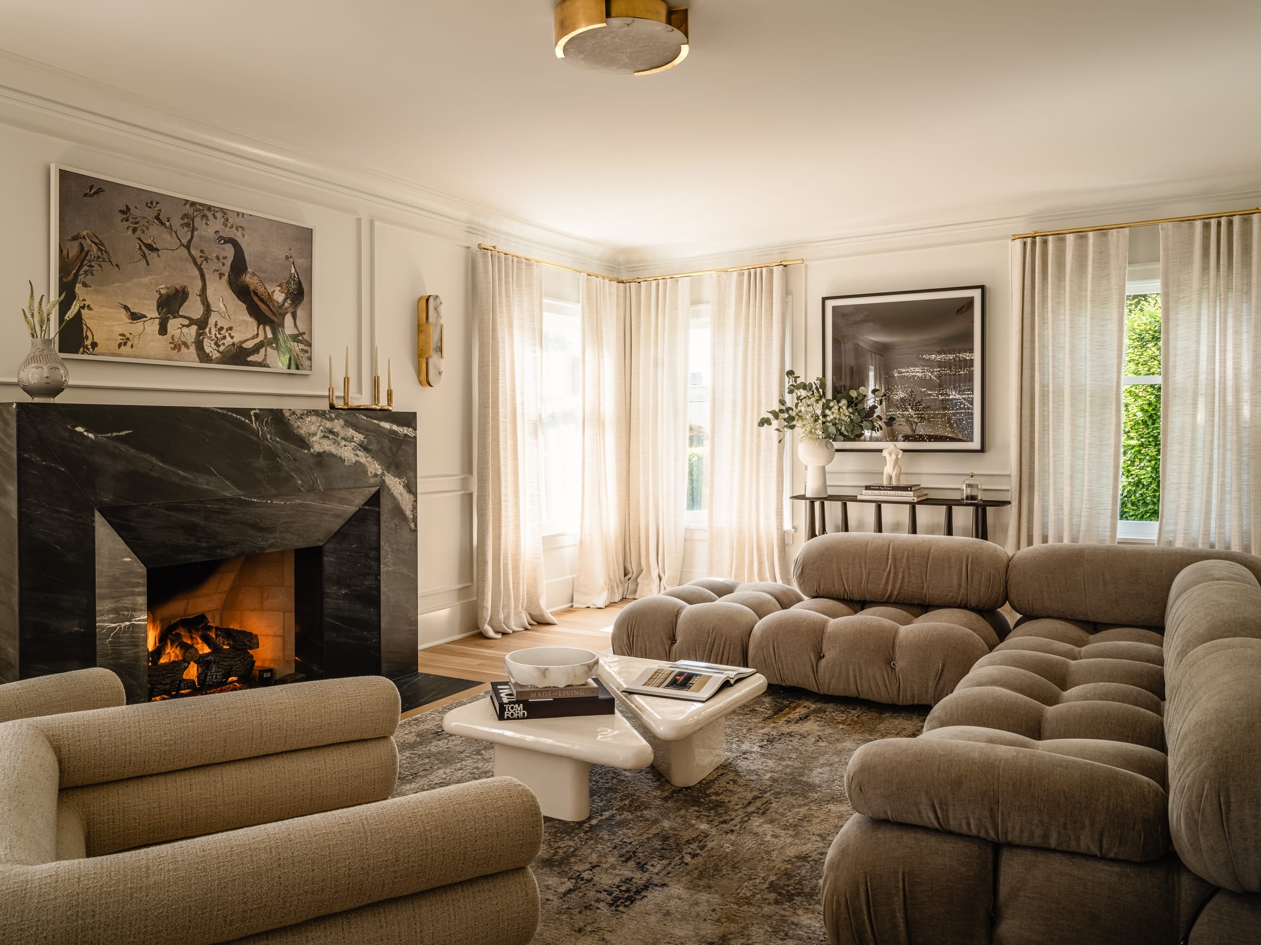 A cozy living room with beige tufted sofas, a black marble fireplace with a painting above, curtains on large windows, and a modern coffee table in the center.