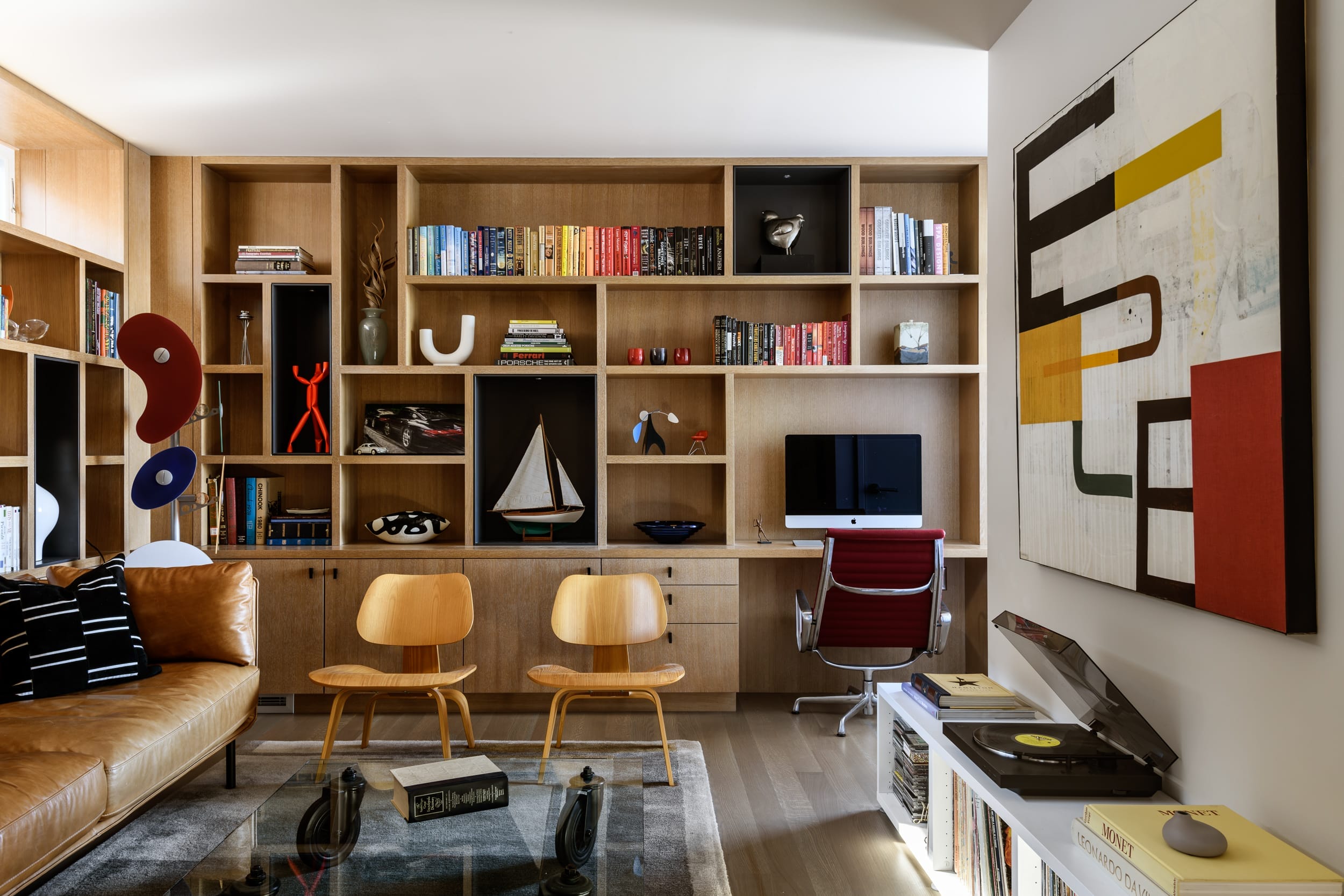 A modern living room with wooden shelves, books, abstract art, a computer, two wooden chairs, a leather sofa, and a turntable.