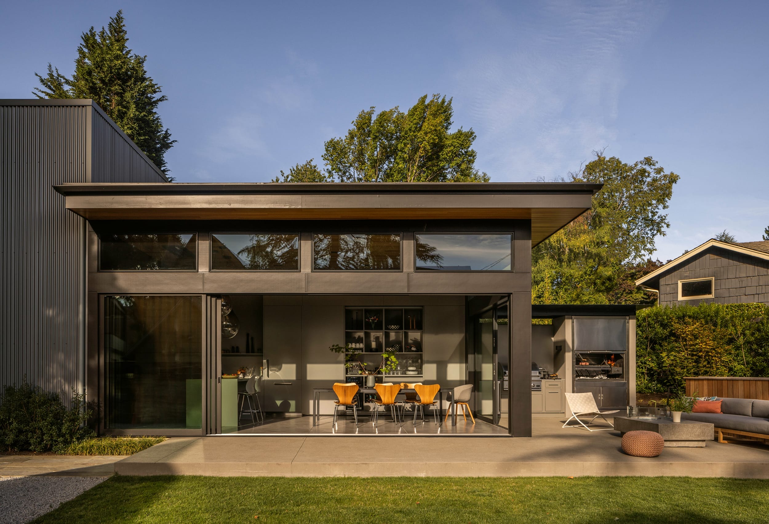 A modern house with large glass windows, a patio with outdoor seating, and a lawn. The dining area is visible inside. Trees and neighboring houses are in the background.