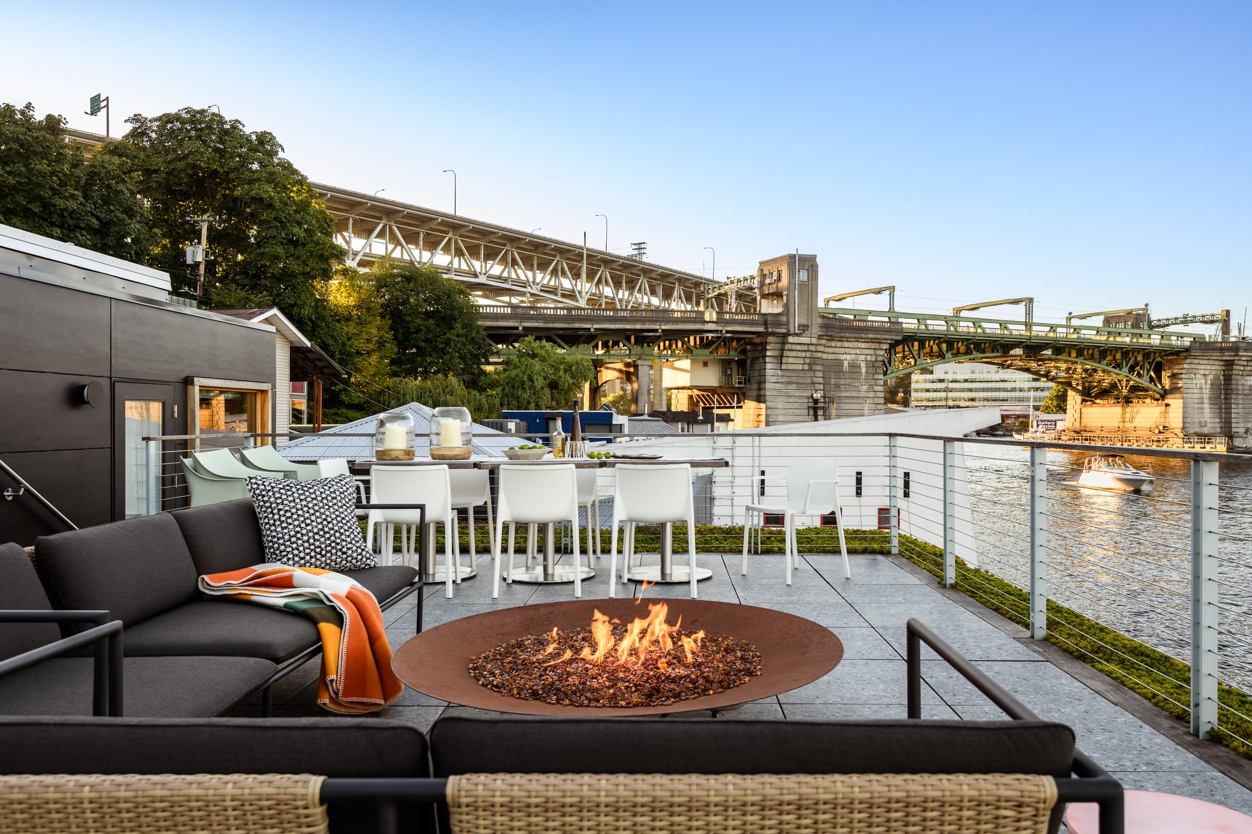 Rooftop patio featuring a fire pit, seating area, and bar stools, overlooking a river and a bridge under a clear blue sky.