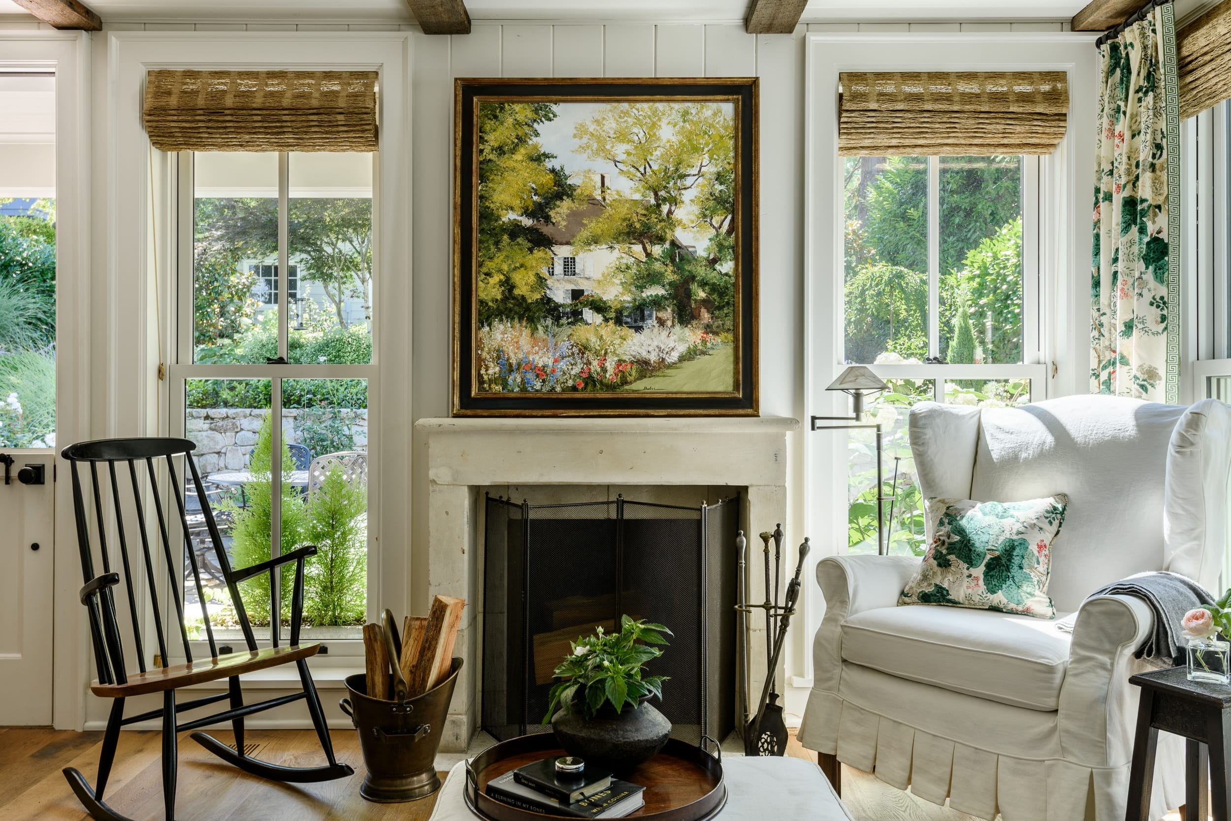 Cozy living room with a fireplace, rocking chair, and armchair. A painting of a garden scene hangs above the fireplace. Windows on either side show a view of greenery outside.