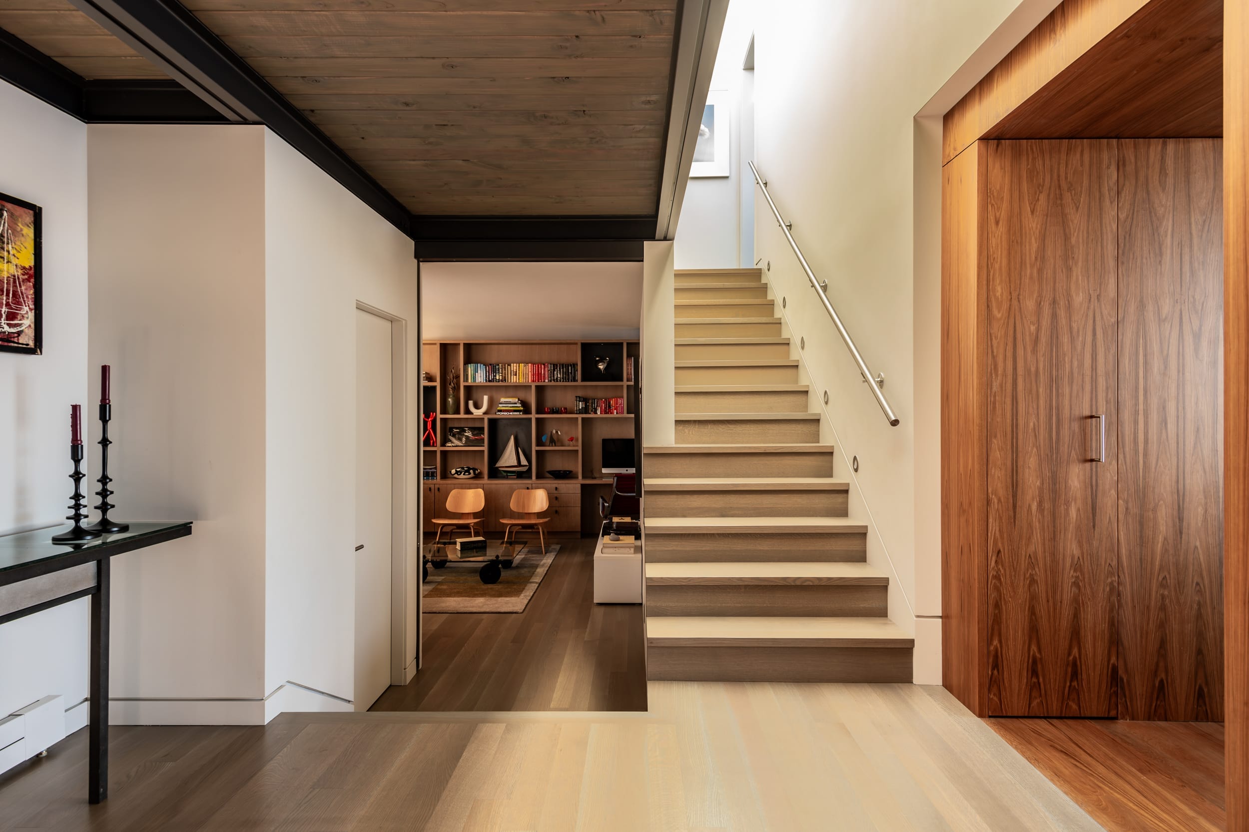 Modern interior with a wooden staircase, a hallway with artwork, and a room with shelves and chairs. Natural light illuminates the space.