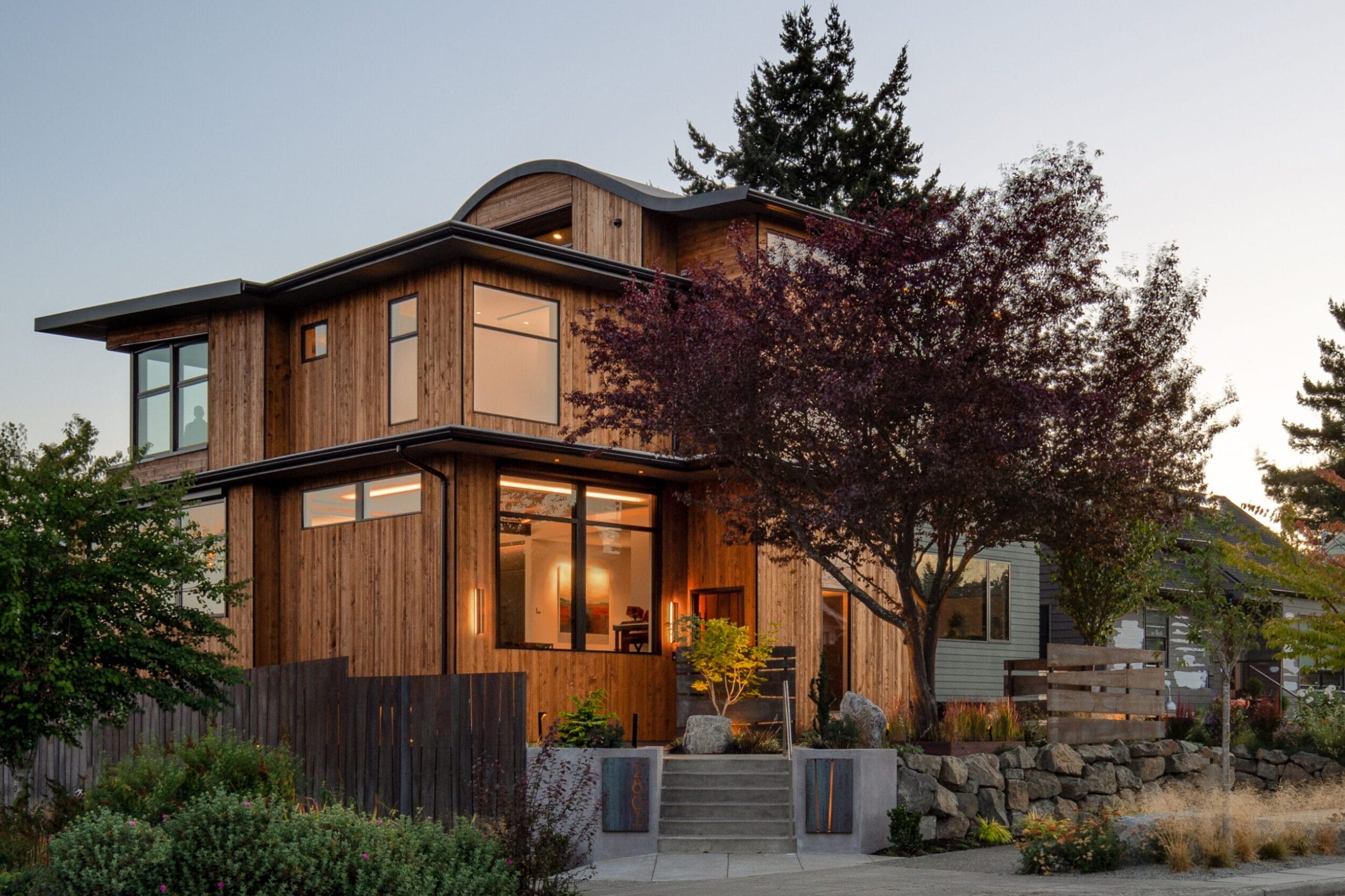A modern wooden house with large windows and a sloped roof, surrounded by trees and shrubs, on a residential street during sunset.
