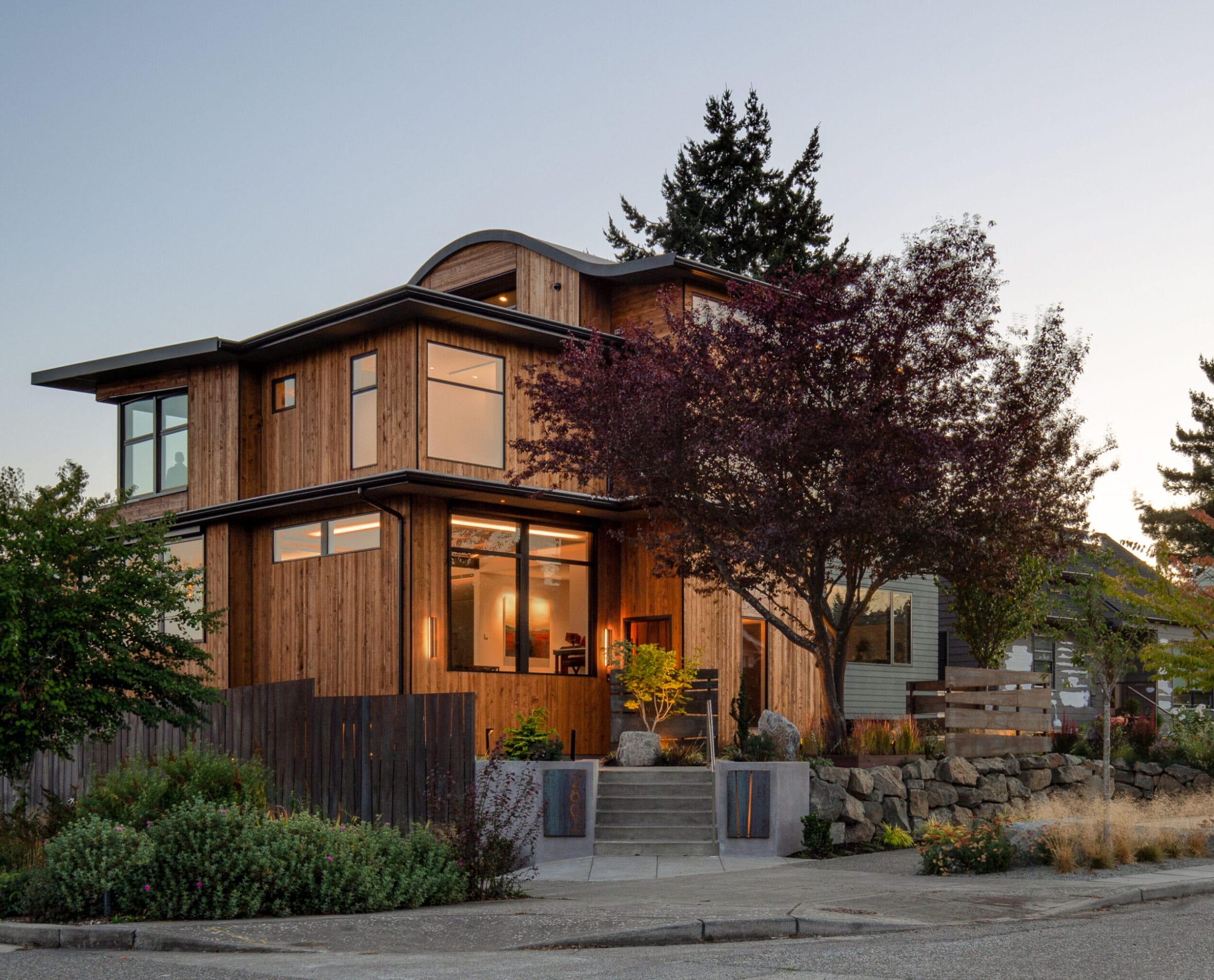 A modern wooden house with large windows and a sloped roof, surrounded by trees and shrubs, on a residential street during sunset.
