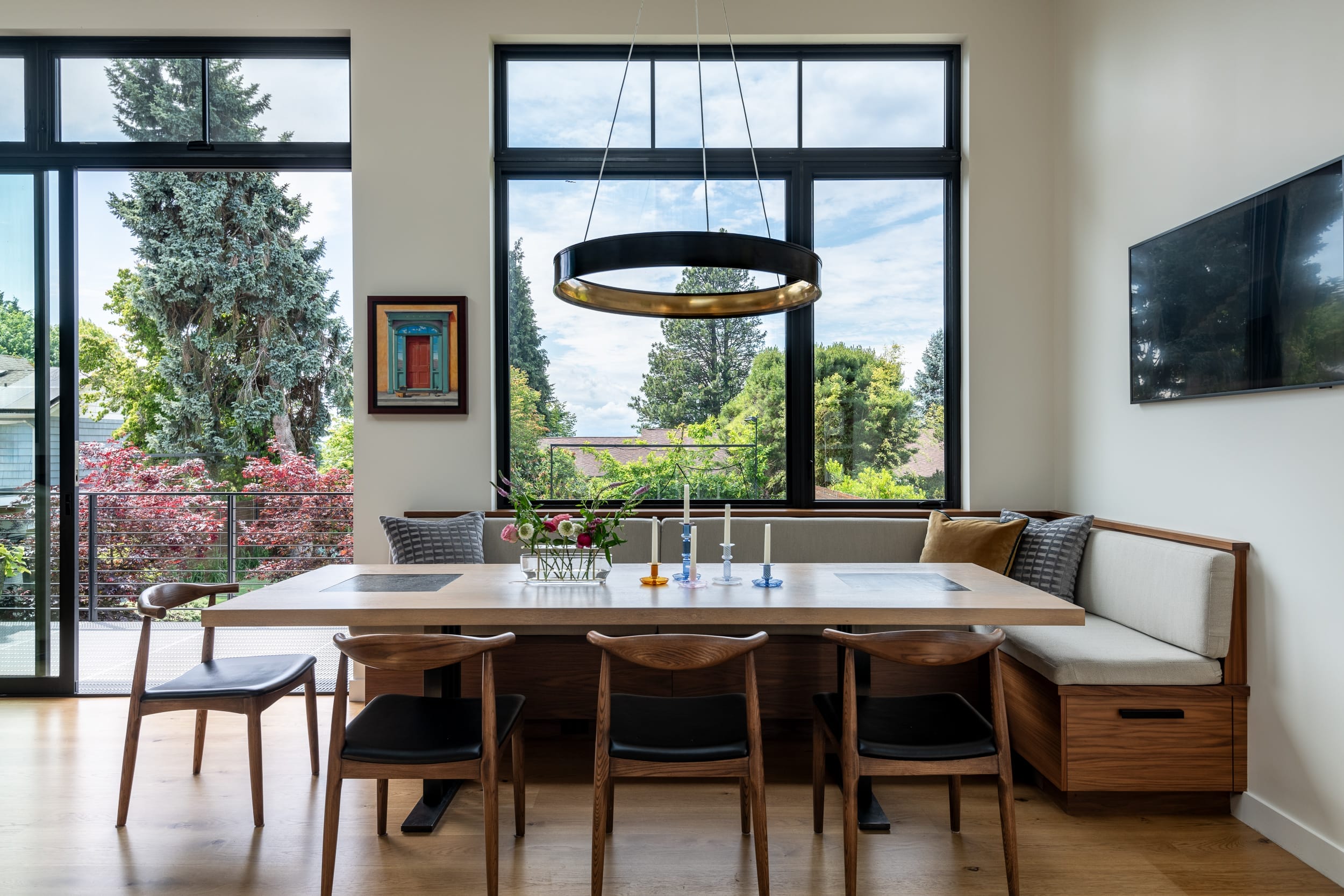 A modern dining room with a large wooden table, six chairs, bench seating, a round ceiling light, large windows, and views of trees and sky. A wall-mounted TV and framed artwork are visible.