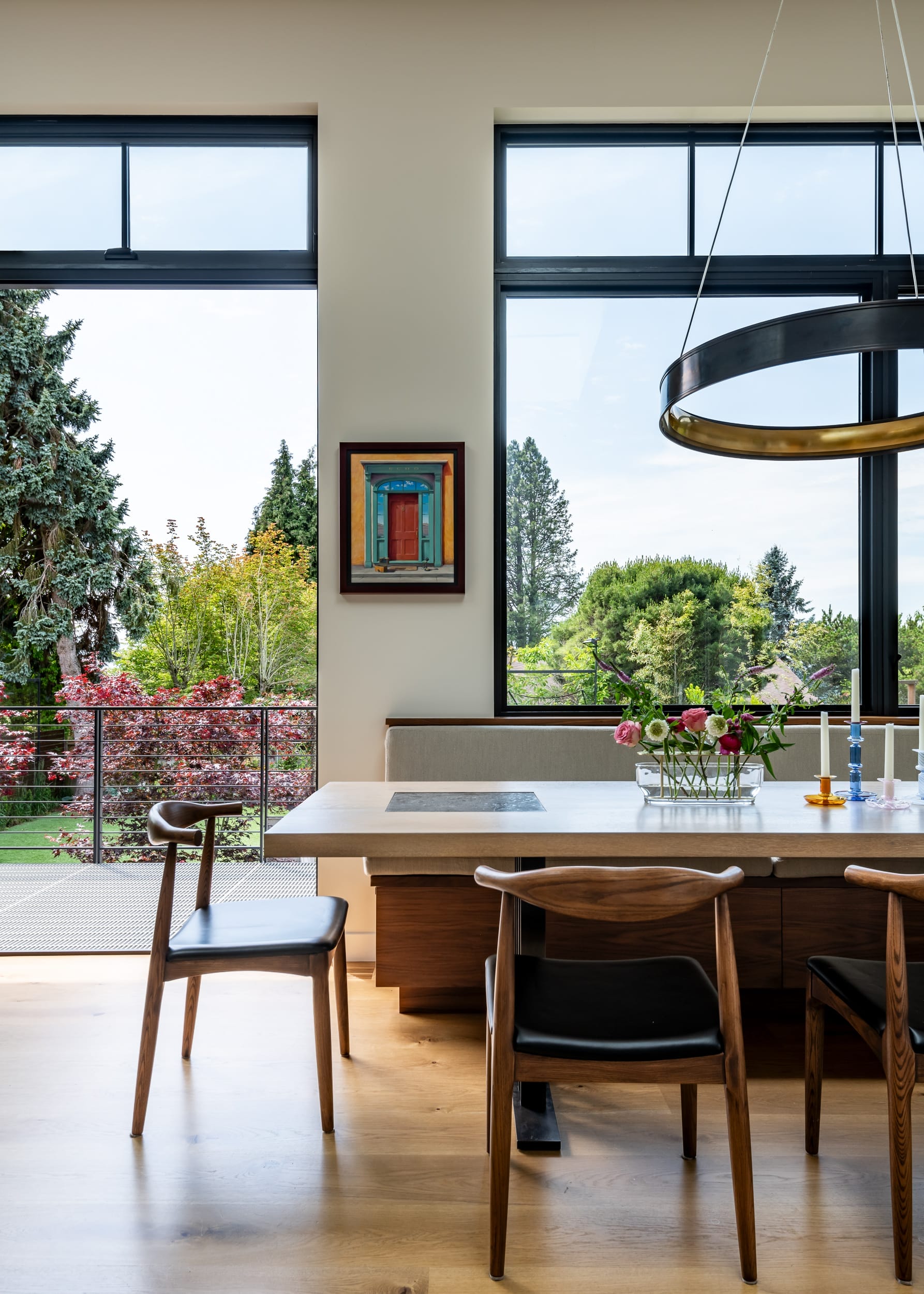 Modern dining room with a wooden table and chairs, large windows showcasing a garden view, a small framed painting on the wall, and a round hanging light fixture above the table.