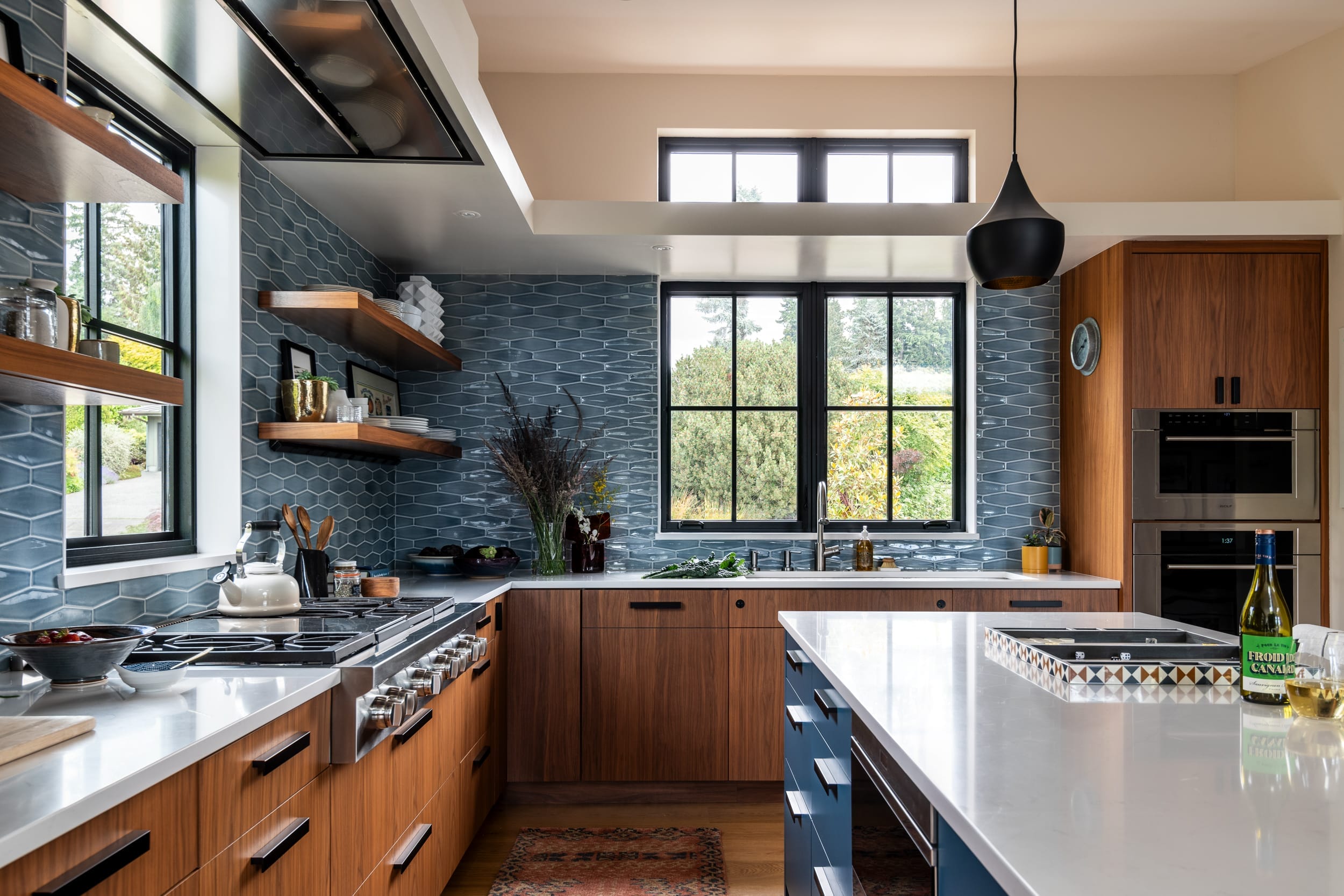 Modern kitchen with wooden cabinets, blue hexagonal tile backsplash, white countertops, and stainless steel appliances. A large central island holds a bottle of wine and glasses.