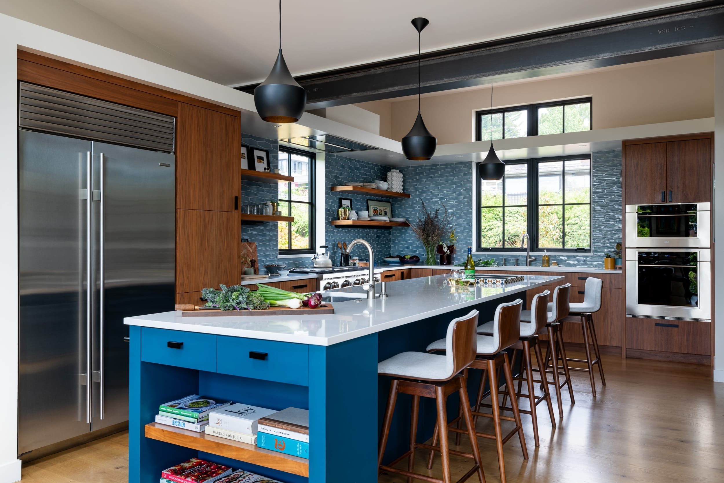 Modern kitchen with a large island, bar stools, blue cabinetry, stainless steel appliances, pendant lights, and open shelving.