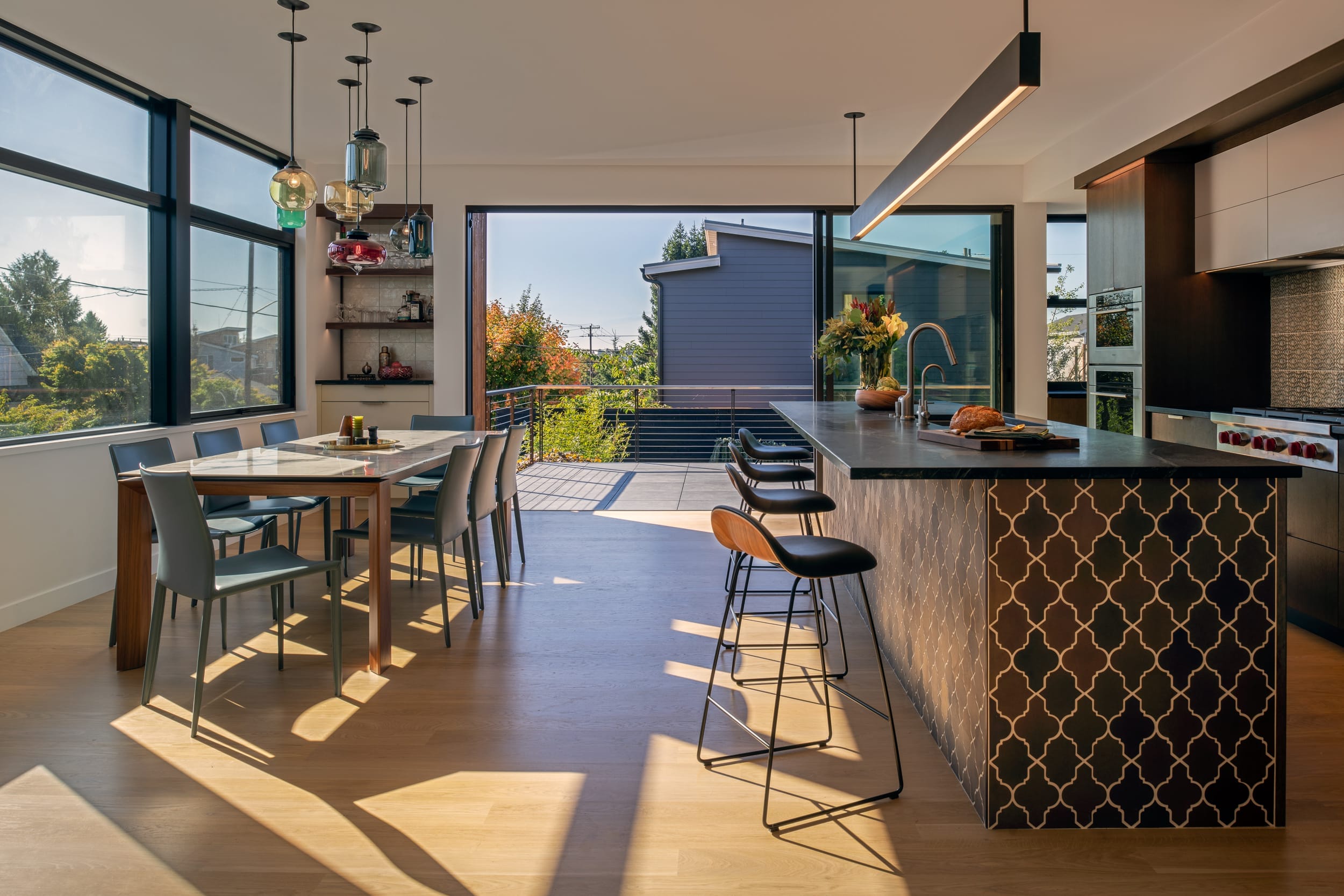 A modern kitchen and dining area with large windows, featuring a wooden table with chairs, an island with barstools, and pendant lights. A view of a patio and trees is visible outside.