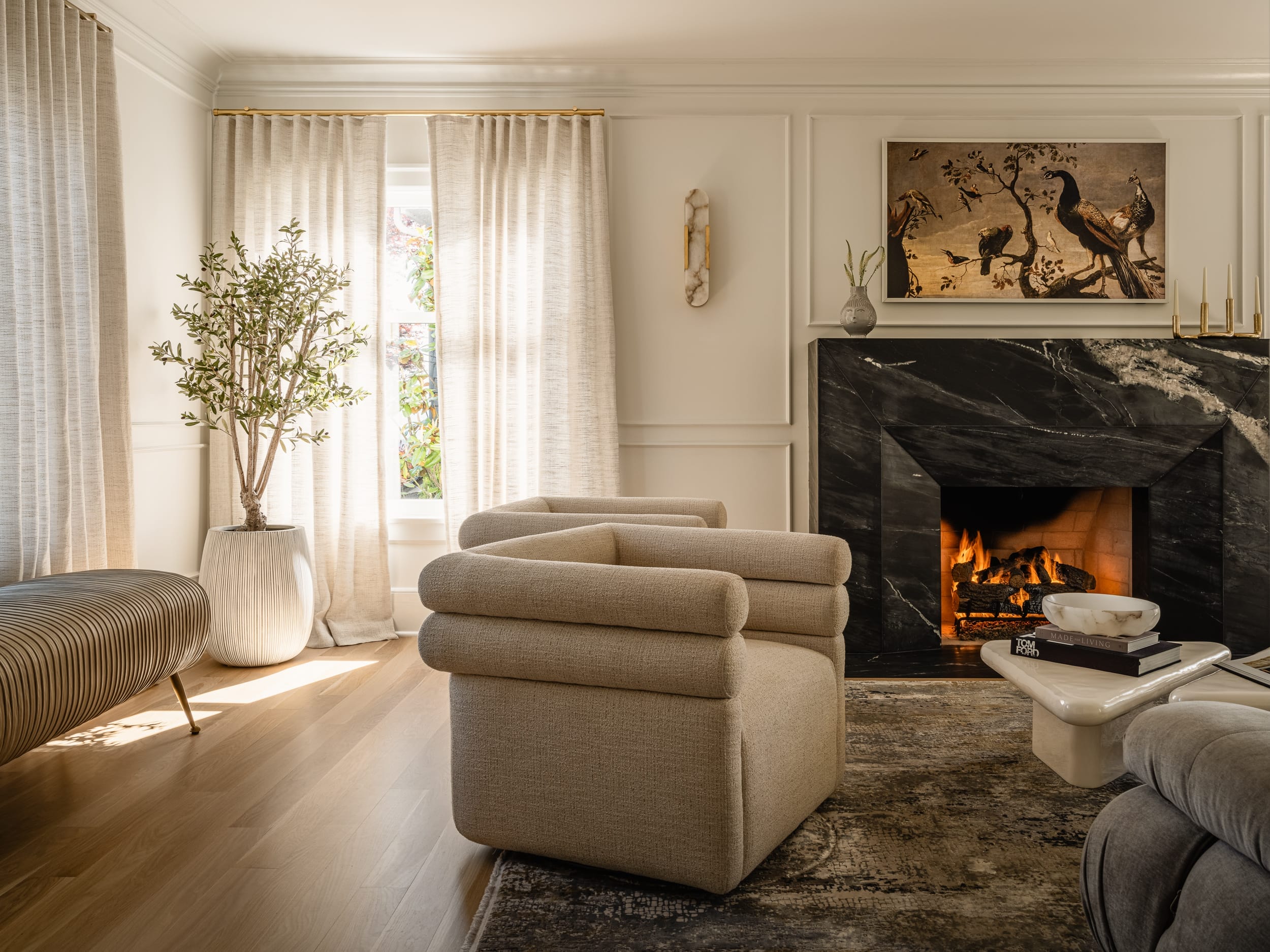 A cozy living room with a lit fireplace, two beige armchairs, a striped bench, and a potted plant. A marble coffee table and a framed painting adorn the space. Natural light filters through sheer curtains.