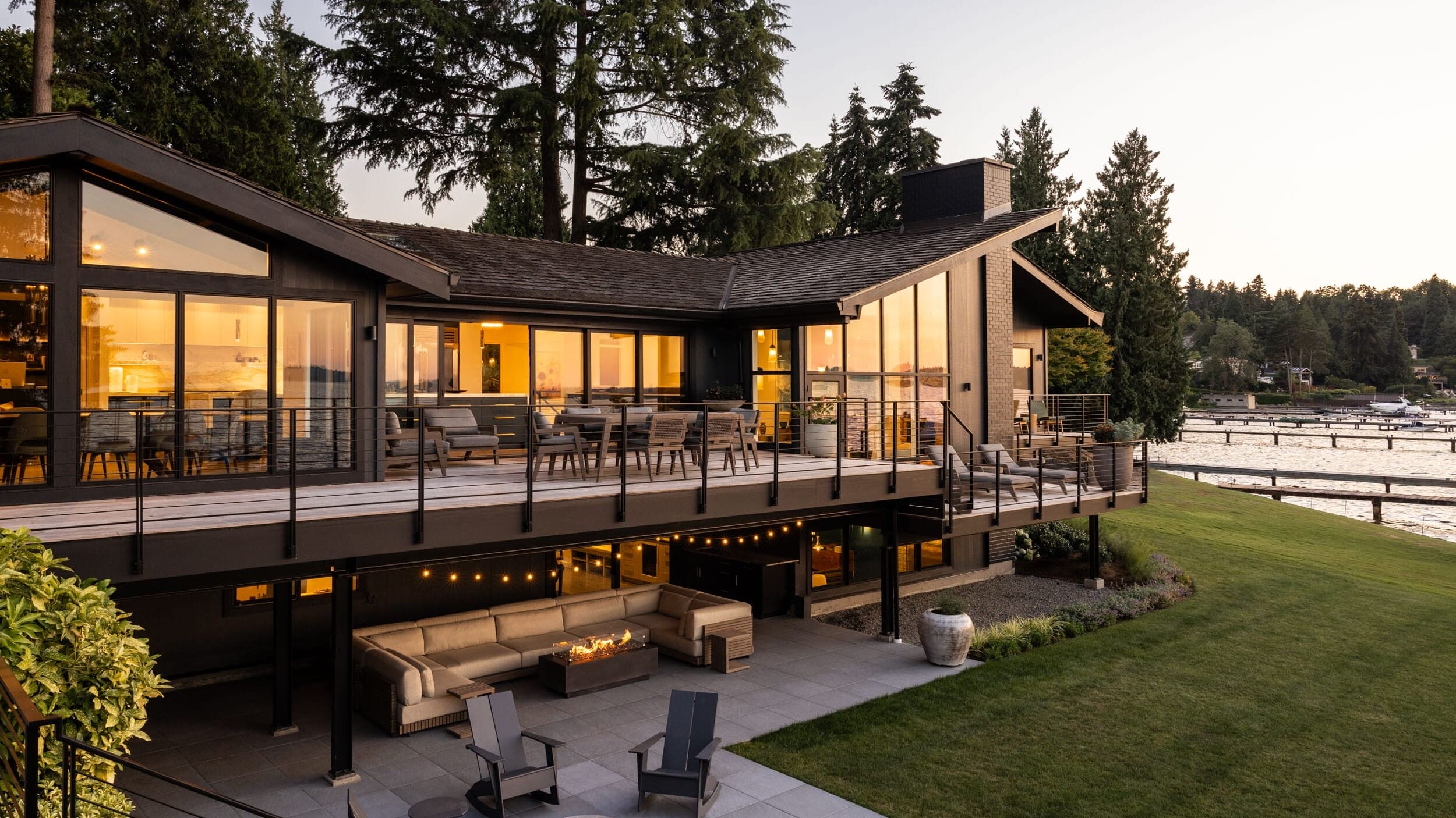 Modern house with large windows and outdoor seating areas on a grassy yard, overlooking a lake with a dock, surrounded by trees at sunset.