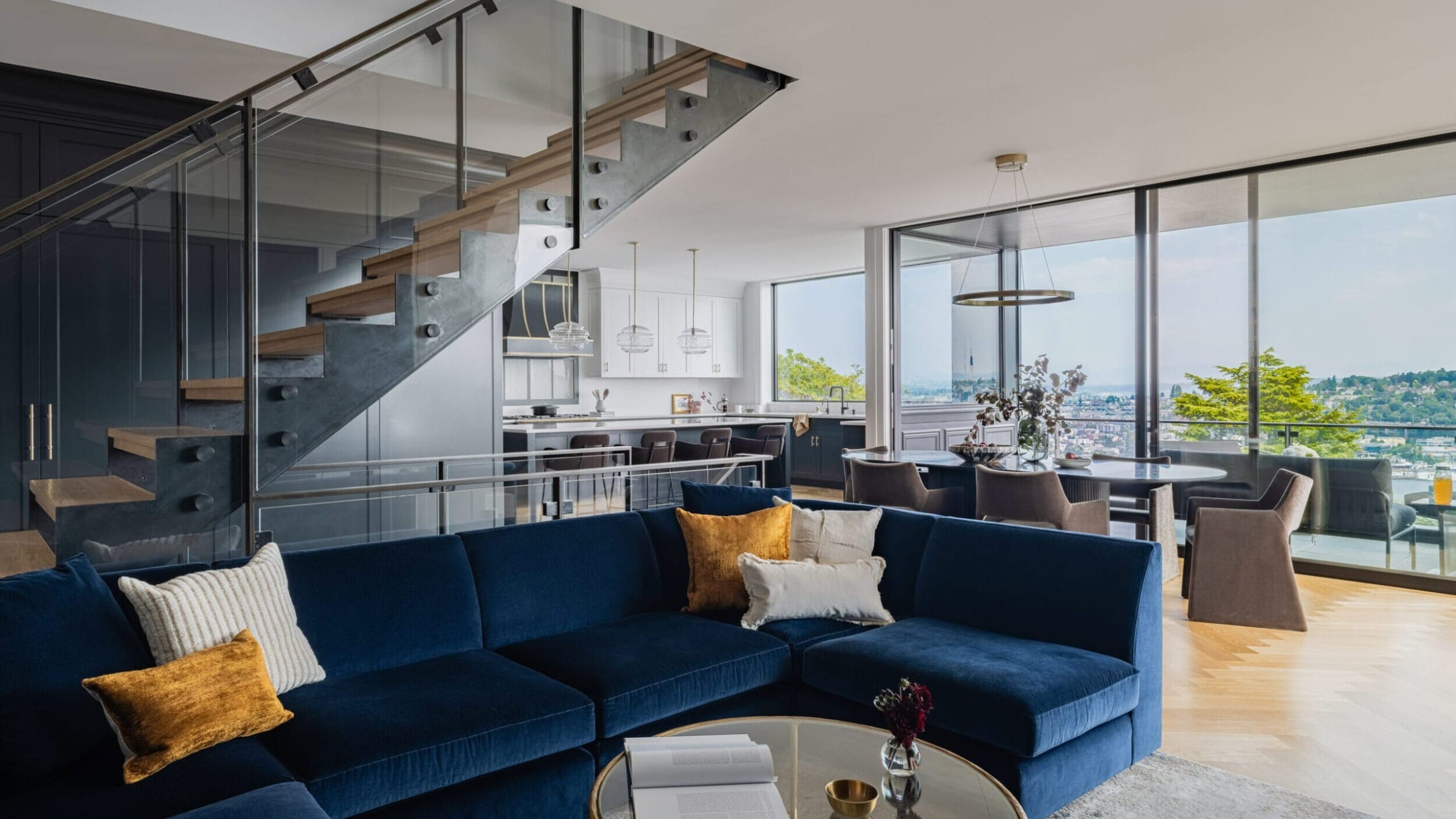 Modern open-plan living room with blue sectional sofa, glass coffee table, and view of a kitchen with island. A staircase with glass railing is on the left, and large windows offer a scenic view.