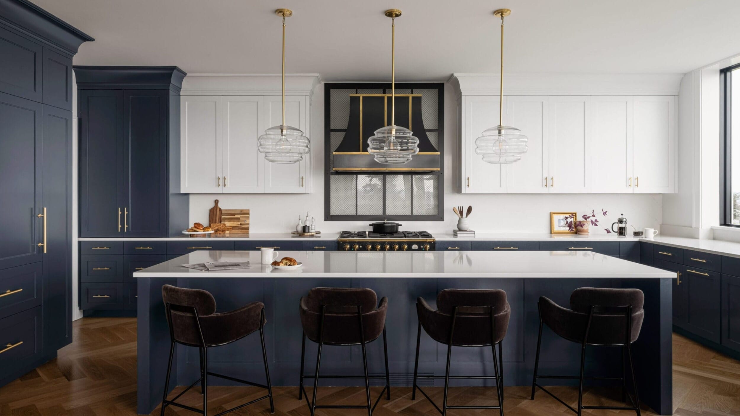 Modern kitchen with a navy island, white cabinets, gold accents, and glass pendant lights. Four dark chairs line the island, and wooden herringbone flooring completes the space.