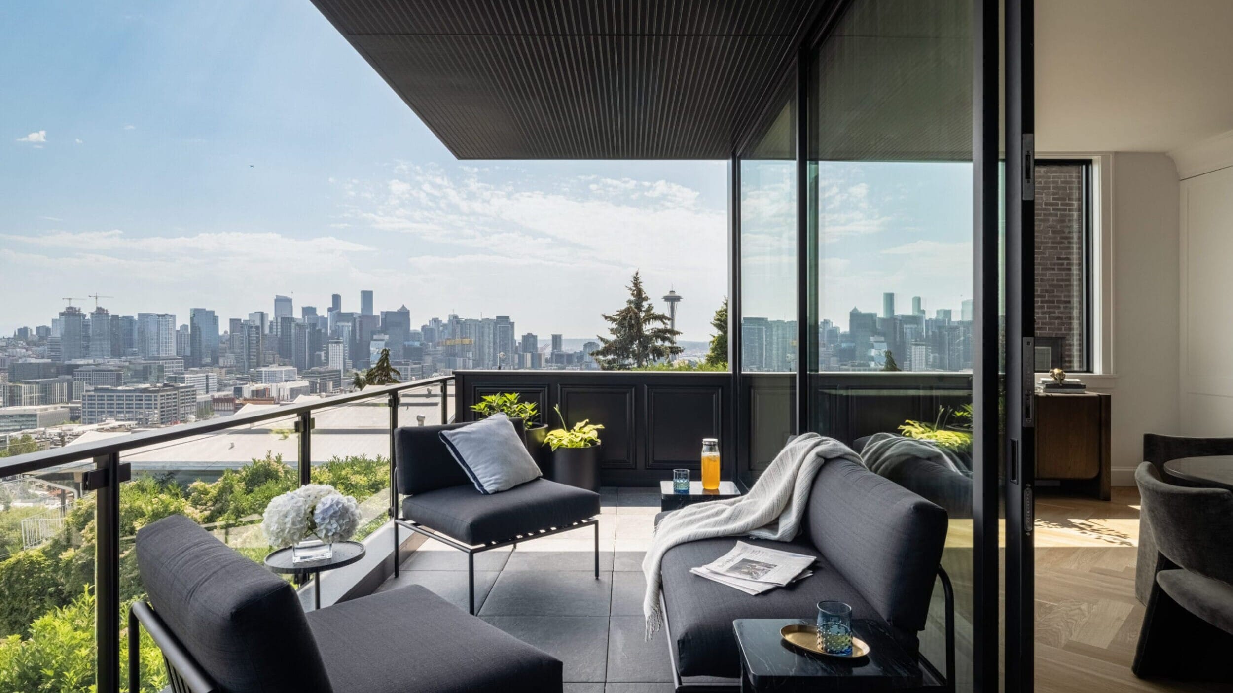 A modern balcony features dark furniture, a glass railing, and a city skyline view in the distance. A blanket and tray with drinks are placed on the couch.