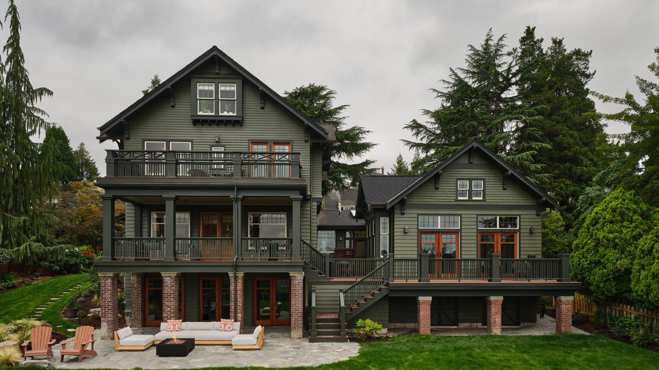 Large green house with three stories, multiple balconies, and a spacious lawn. Surrounded by trees, with outdoor seating on a stone patio.