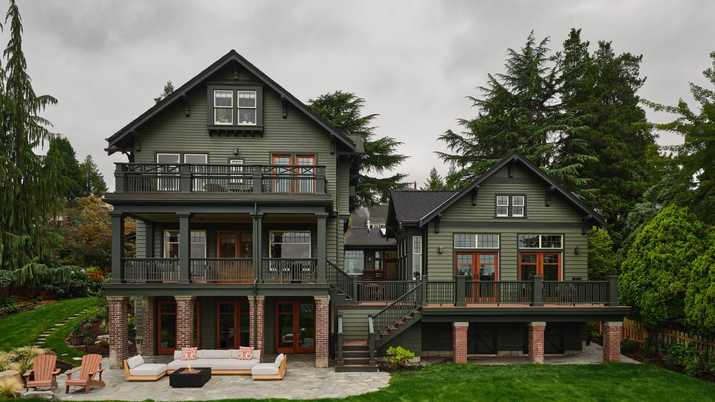 Large green house with multiple balconies and a patio in a wooded area, featuring outdoor furniture on a stone terrace and surrounded by lush greenery.