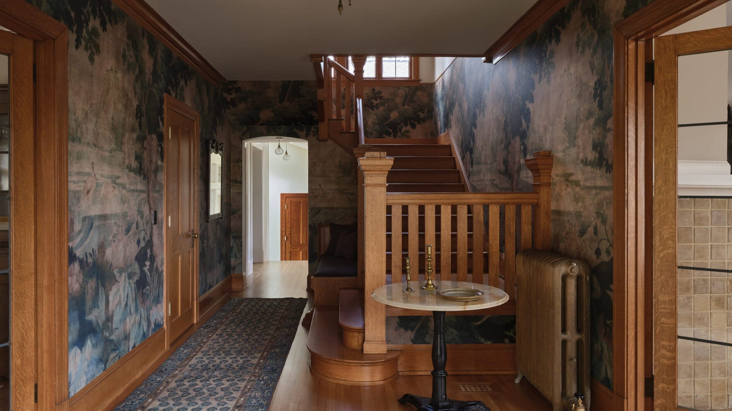 Vintage hallway with wooden floors, ornate wallpaper, and a staircase. A small table and chairs are against the wall. A patterned rug leads towards an open doorway.