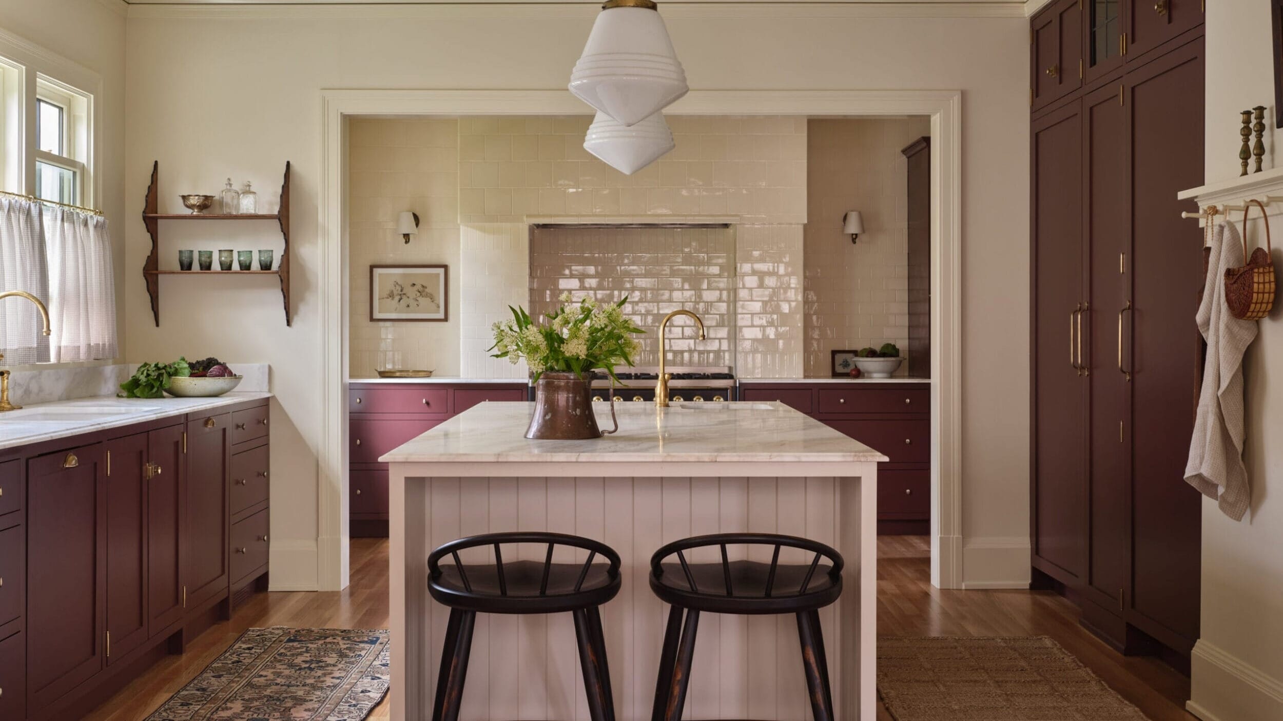 A kitchen with a central island, two black stools, burgundy cabinets, and a white pendant light. There are wooden floors, a rug, and green plants in a vase on the island.