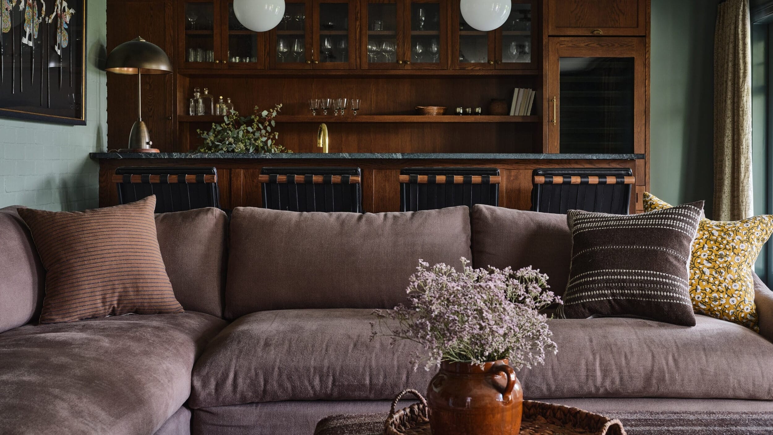 A cozy living room with a large gray sectional sofa, decorative pillows, and a coffee table with a vase of flowers. A wooden kitchen bar with cabinets and pendant lights is in the background.