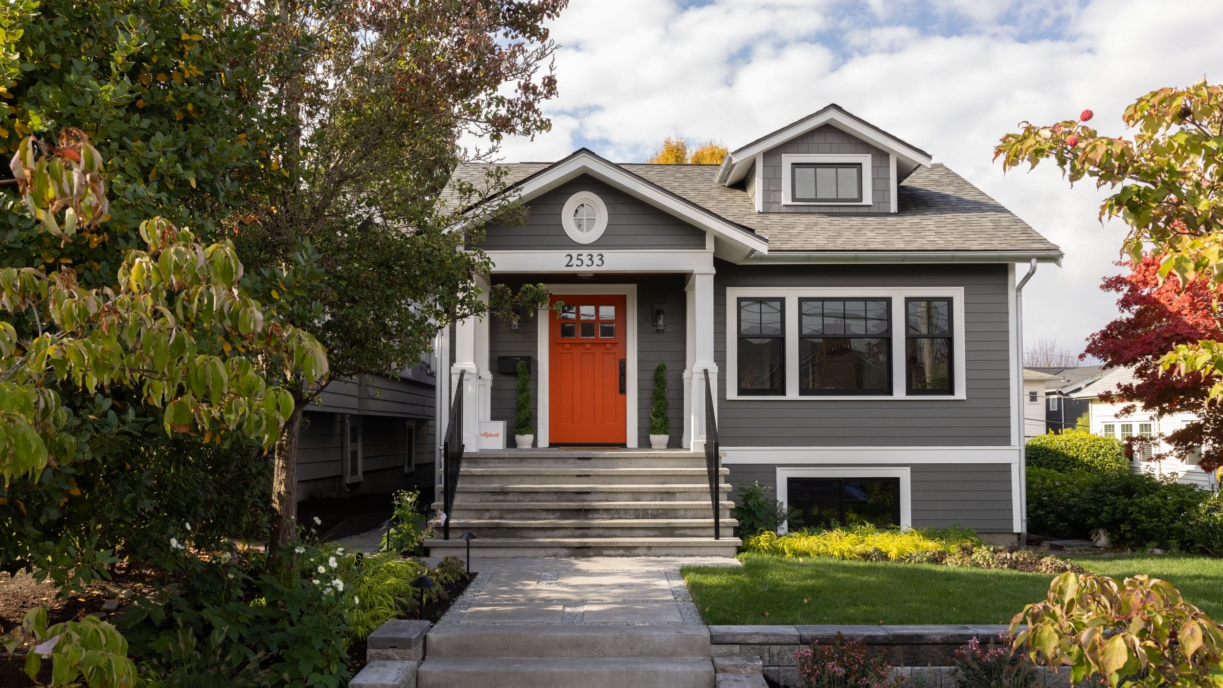 A gray house with a vibrant orange door, surrounded by trees and shrubs. Steps lead up to the porch, and the house number 