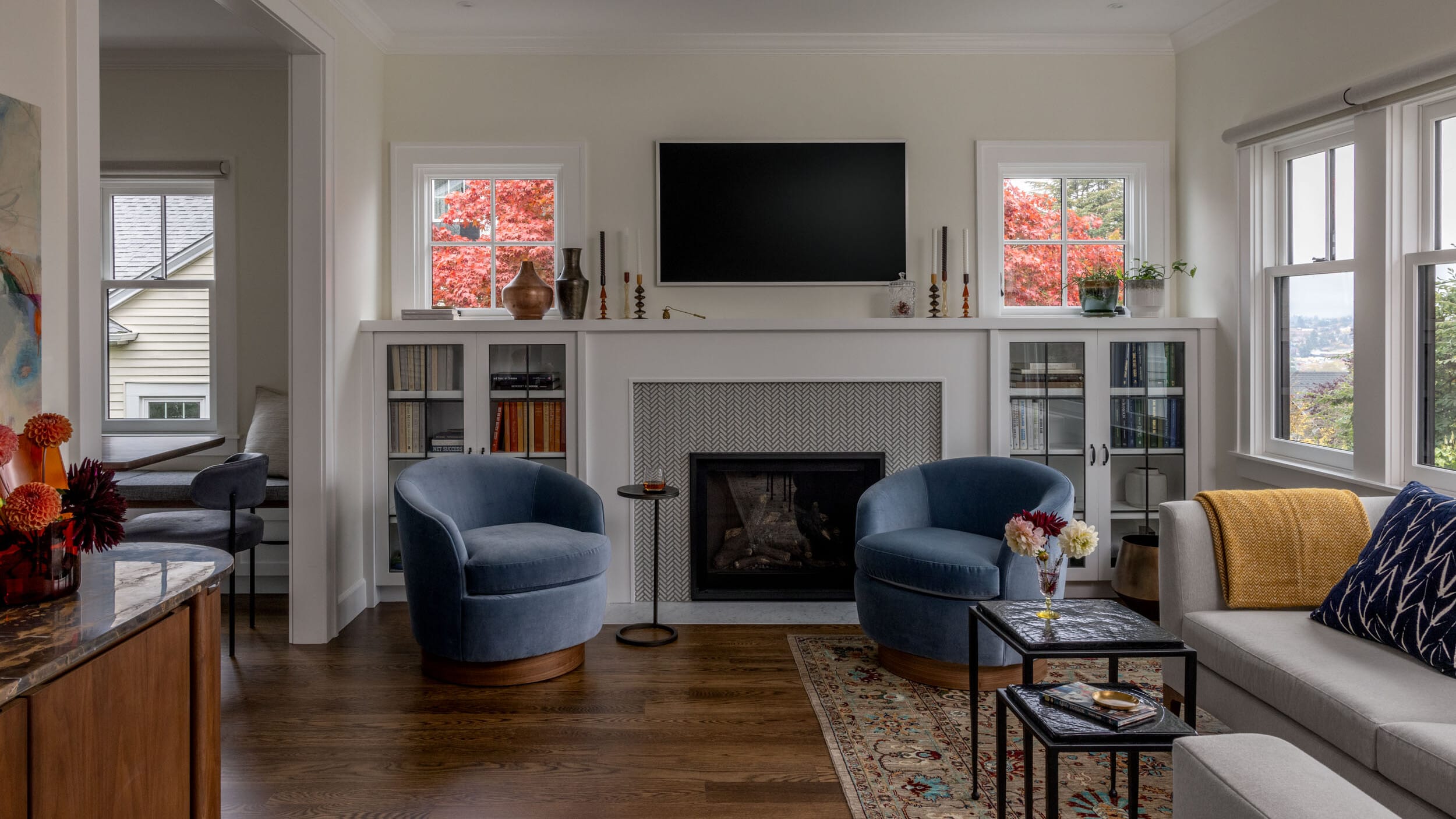 Cozy living room with a fireplace, two blue armchairs, a gray sofa, and a mounted TV. Large windows show red autumn foliage outside. A patterned rug and plants add warmth.