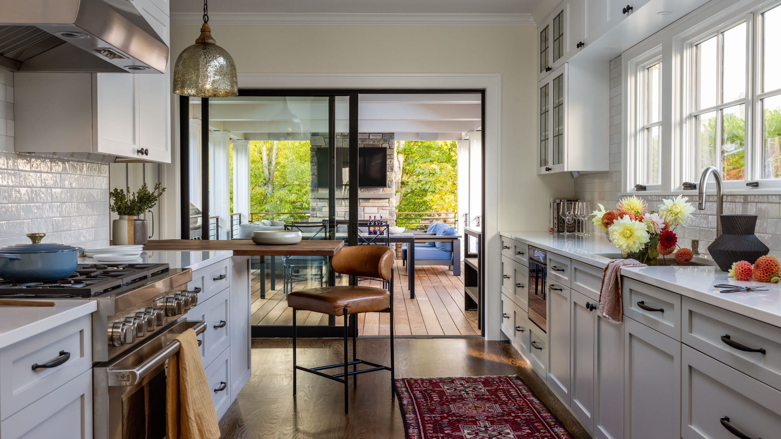 A modern kitchen with white cabinets, stainless steel appliances, and a red rug. There's a bar stool and a glass door leading to a patio with outdoor seating.