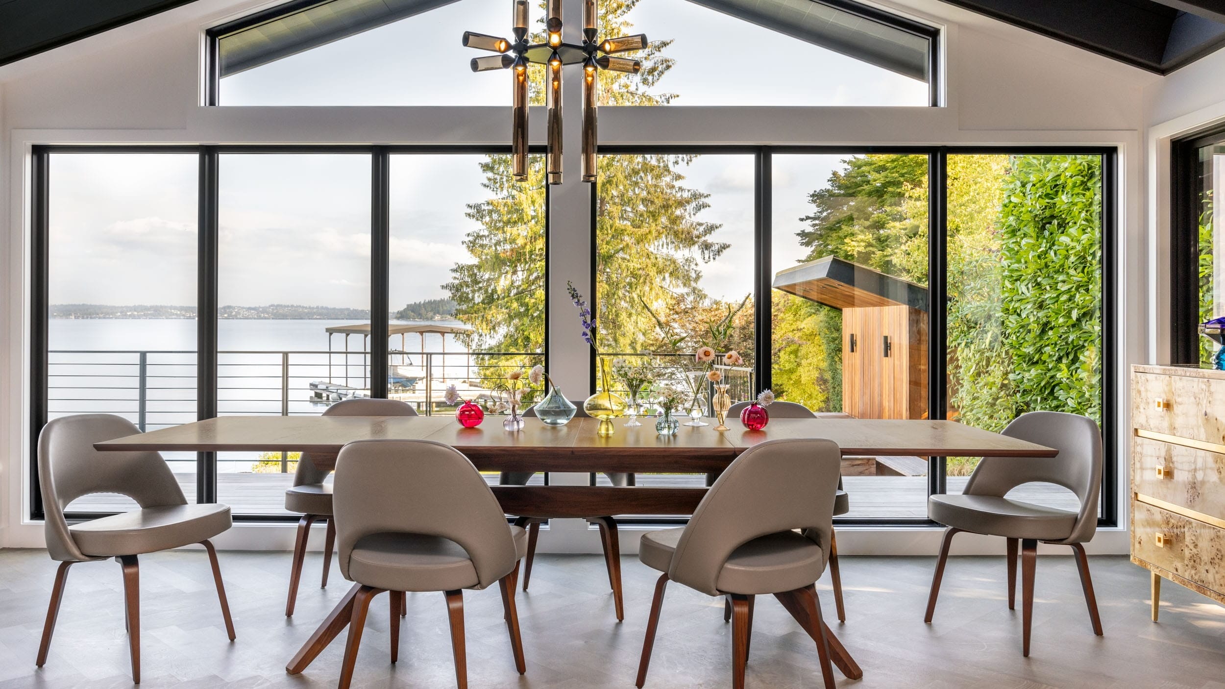 A modern dining room with a wooden table, six chairs, and a geometric chandelier. Large windows offer a view of trees and a lake.