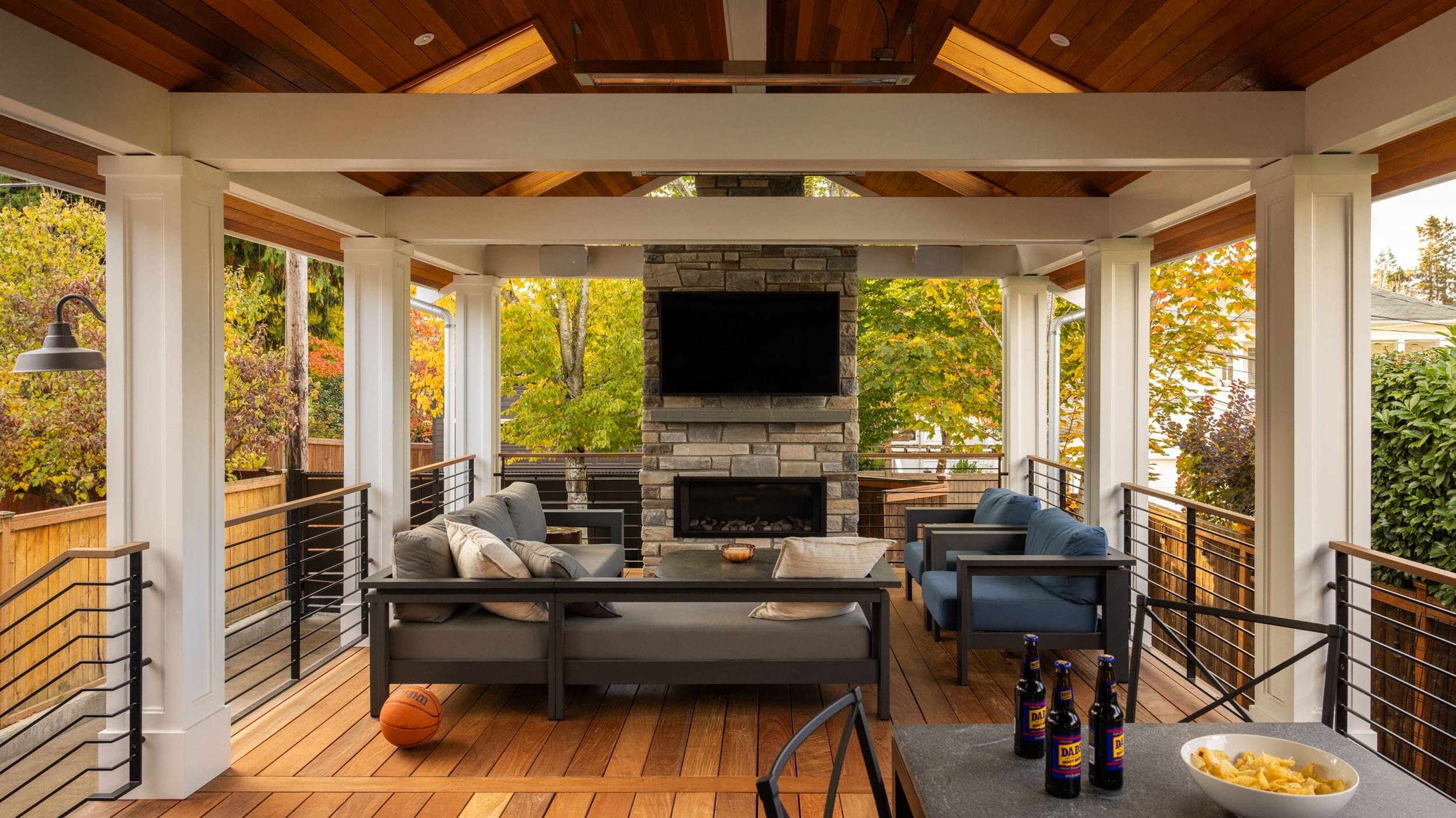 Covered outdoor seating area with a TV above a stone fireplace, surrounded by gray sofas and a wooden floor. A basketball, bottles, and a bowl of snacks are visible on a table.