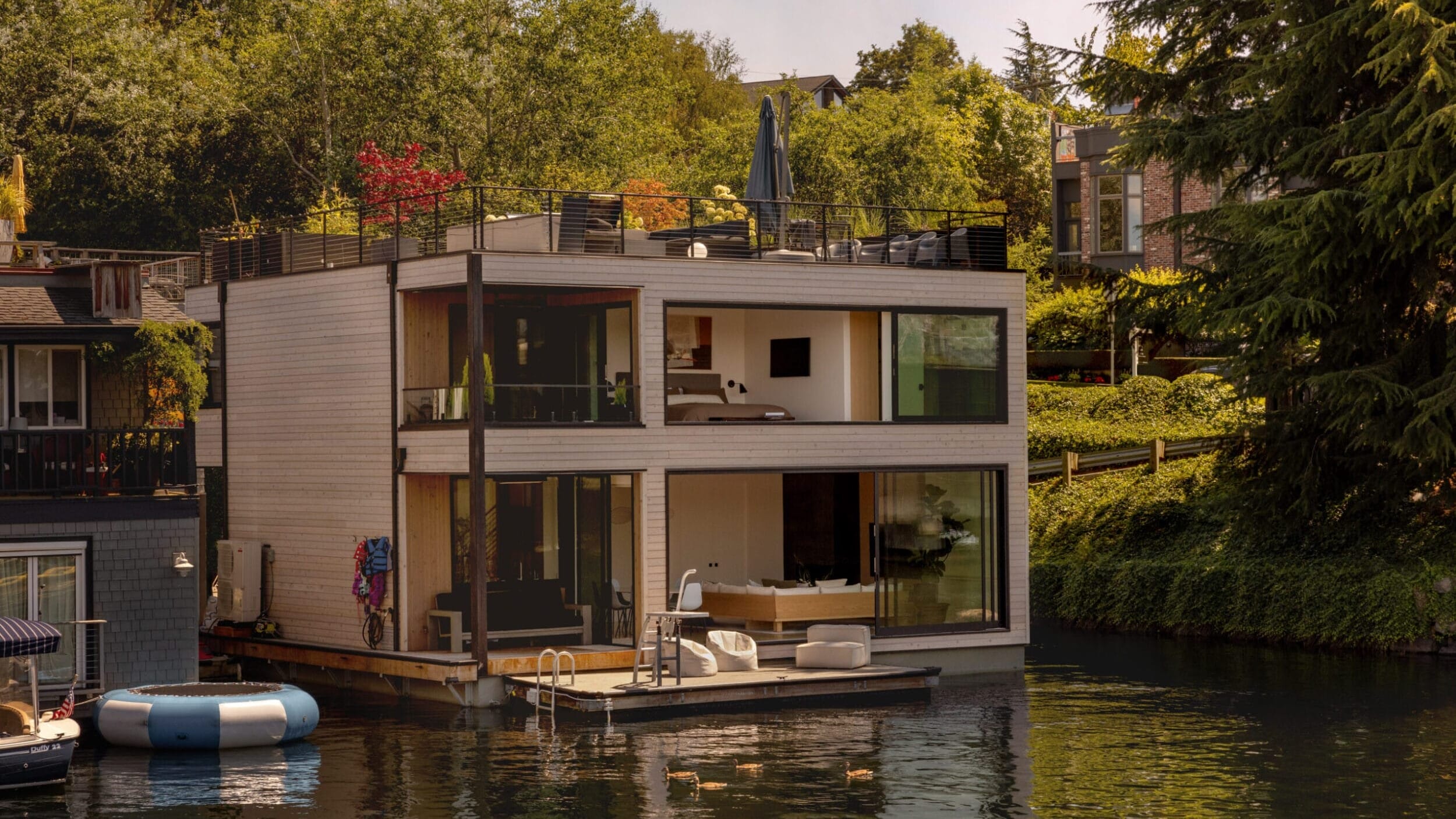 A modern, two-story floating house with large glass windows and a rooftop deck is situated on a calm lake, surrounded by greenery.