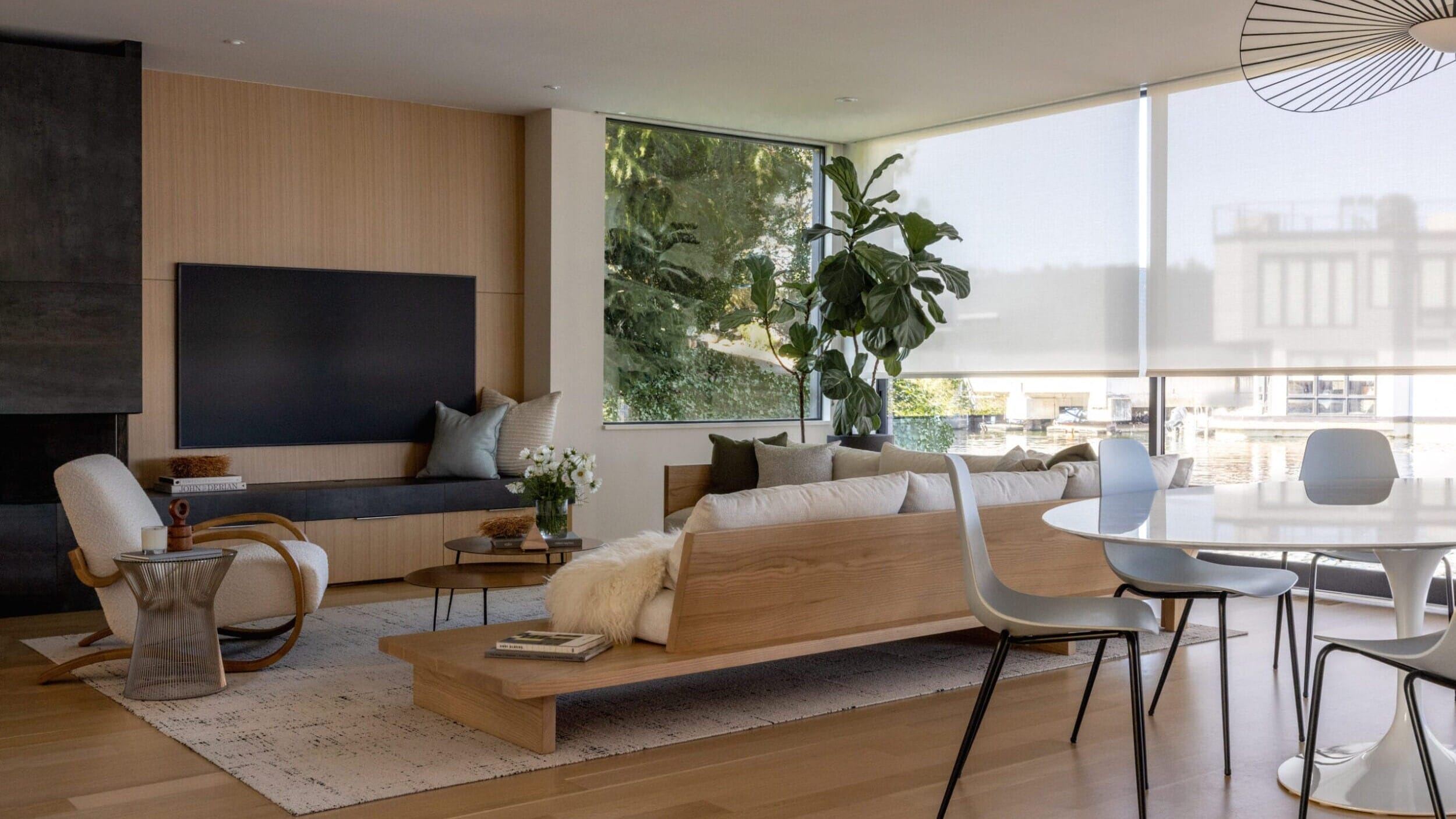 Modern living room with a wooden floor, contemporary furniture, a large window with blinds, a plant, and a dining table with chairs.