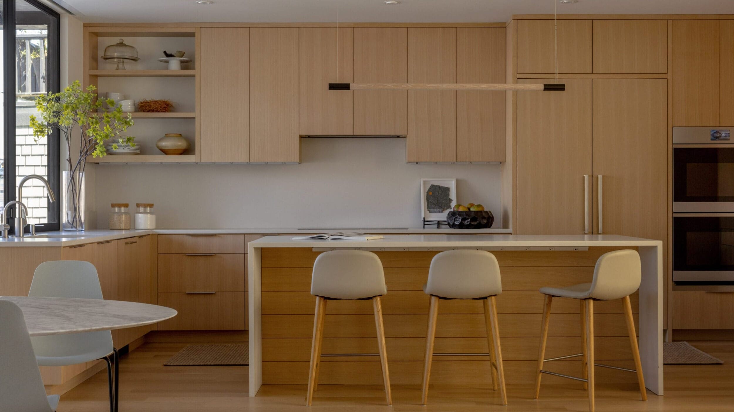 Modern kitchen with light wood cabinets, a large island with three stools, built-in appliances, and a dining area with a round table.