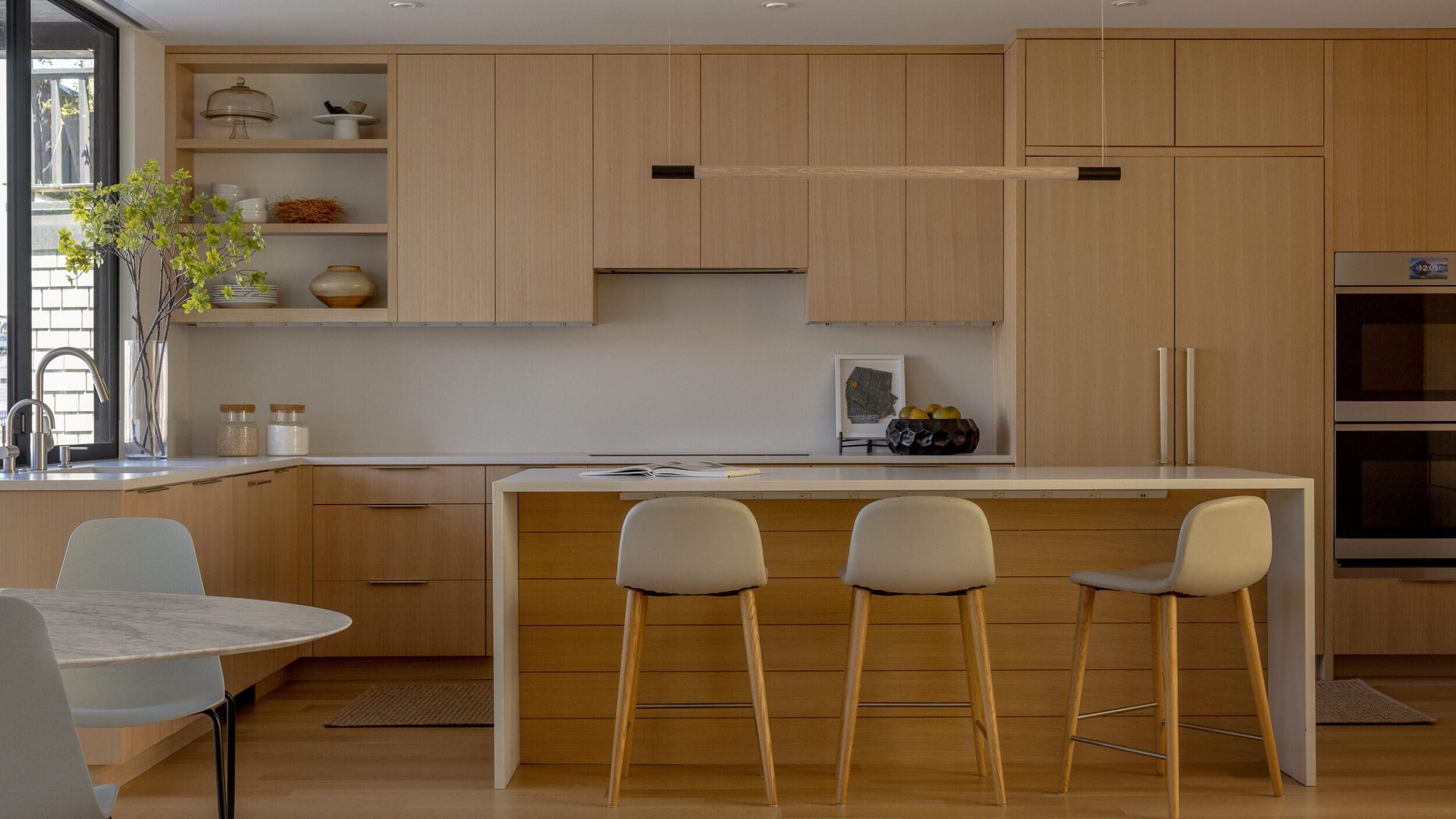 Modern kitchen with light wood cabinets, a central island with three gray stools, and a round dining table. Large window provides natural light. Built-in oven and decor items present.