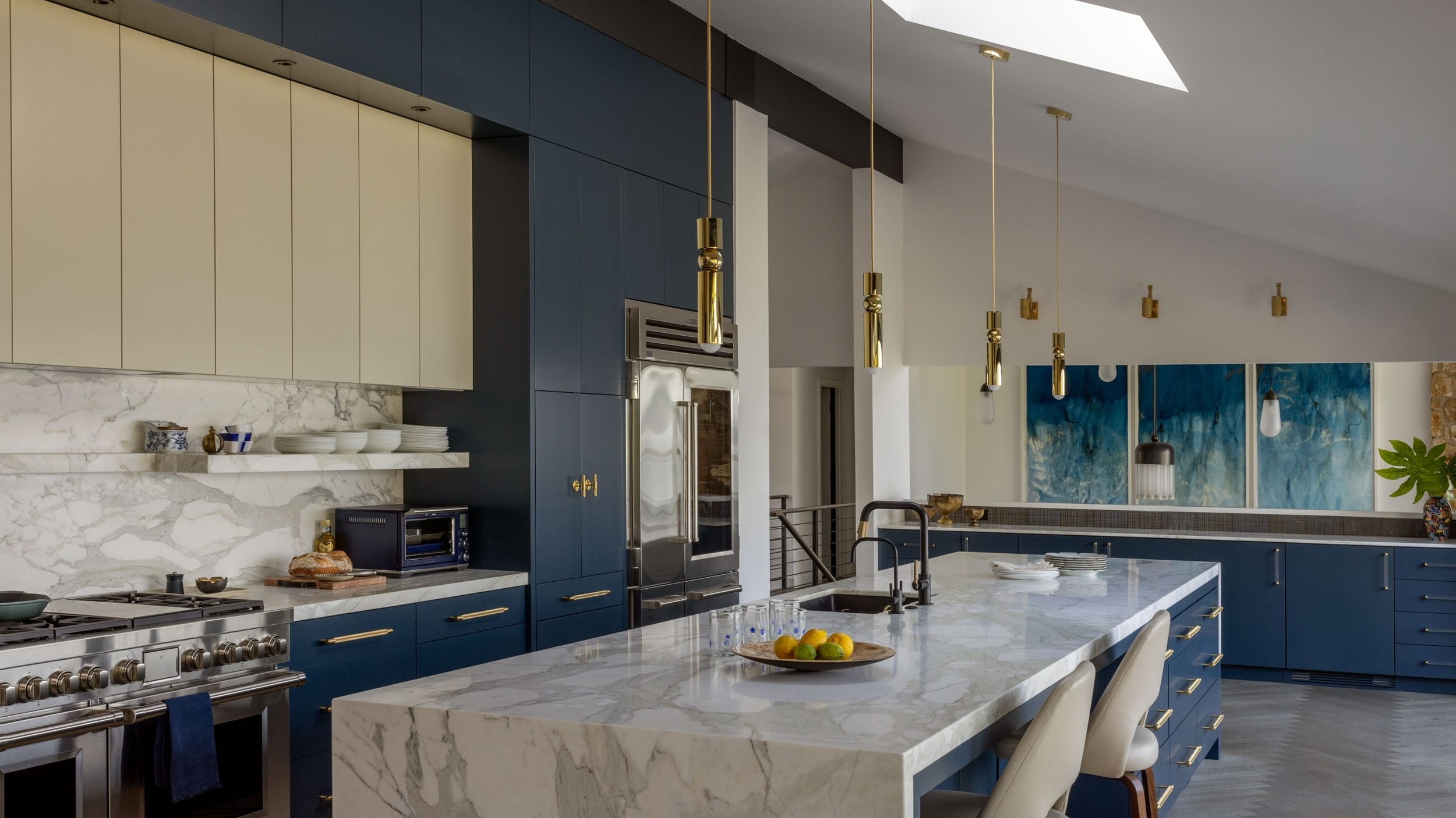 Modern kitchen with marble countertops, navy cabinets, gold accents, and skylight. Island features bar stools and a fruit bowl. Art pieces hang on the wall.