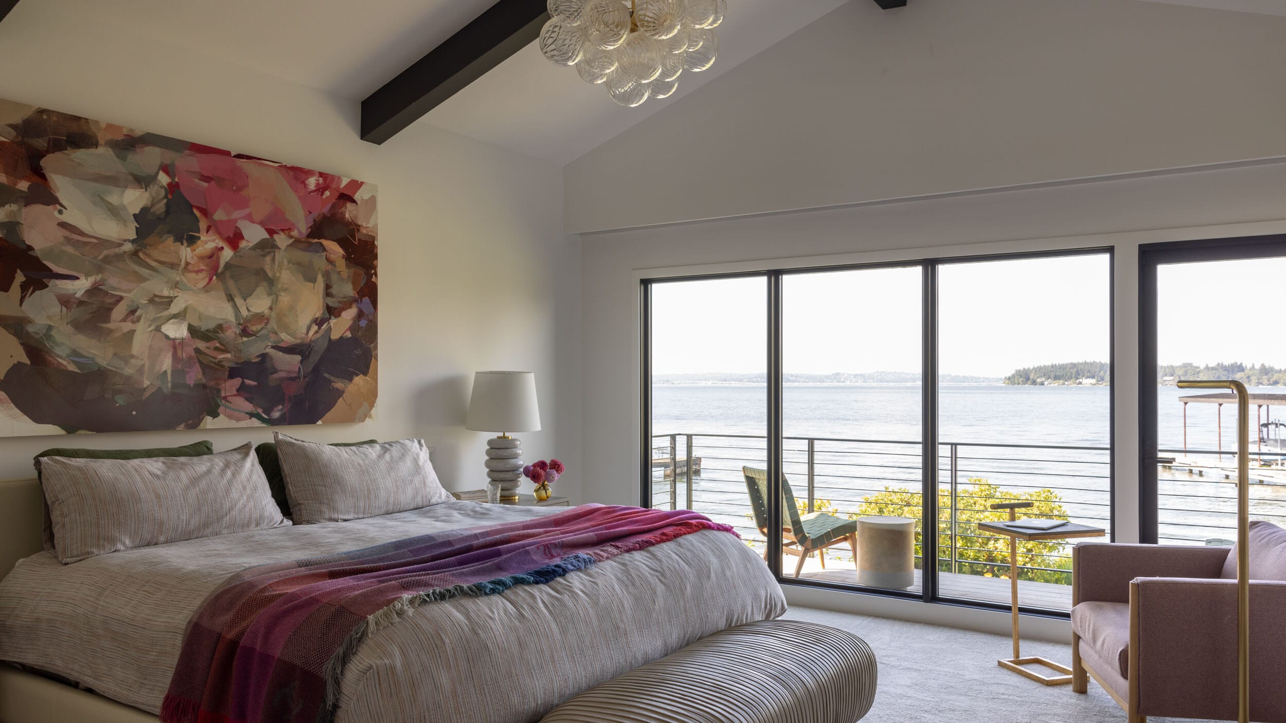 Modern bedroom with large windows overlooking a lake, featuring a bed with a colorful throw, a pink chair, a lamp, and abstract artwork on the wall.