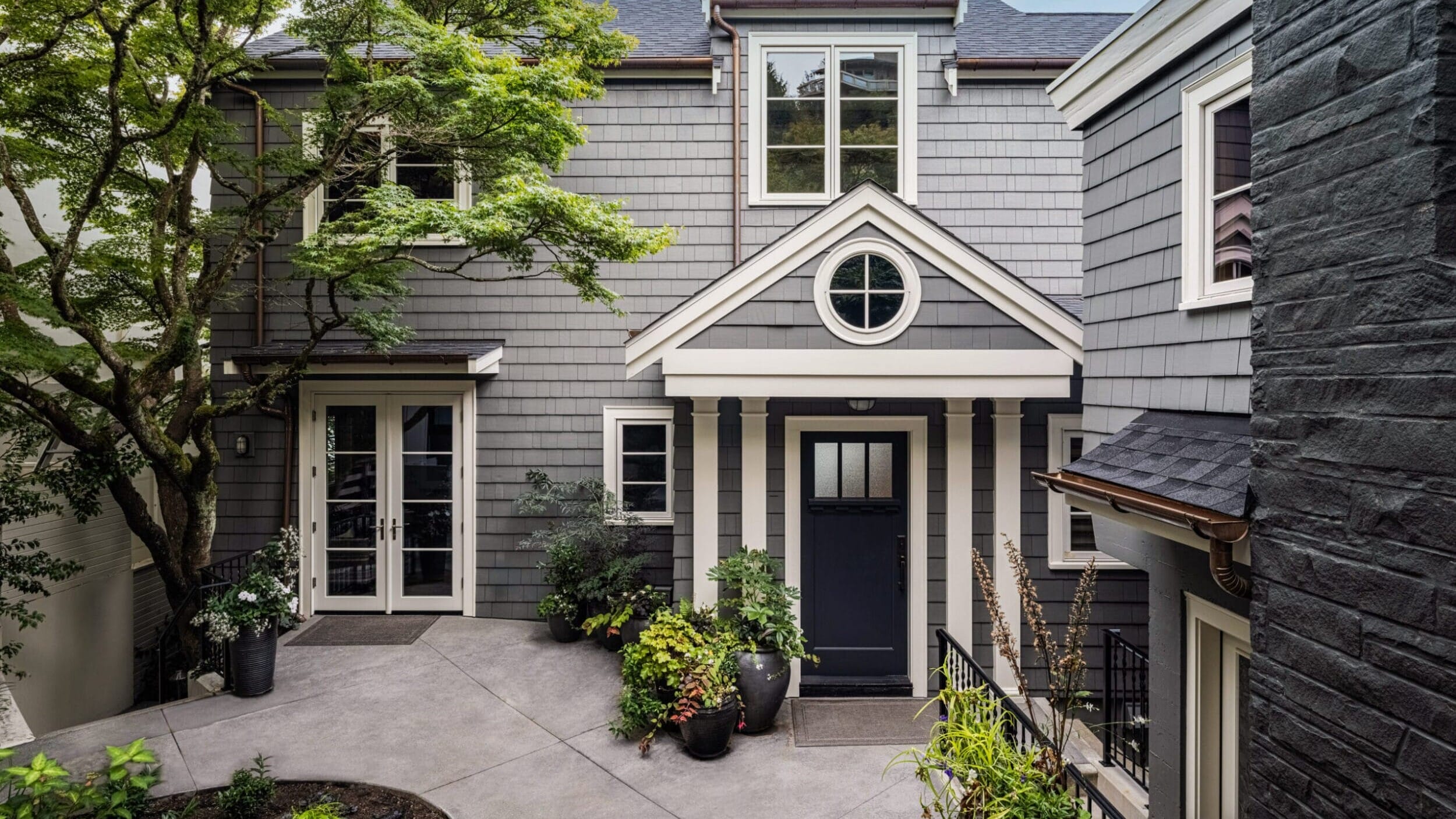 Gray two-story house with a central black door, circular window above, surrounded by greenery and potted plants on a concrete courtyard.