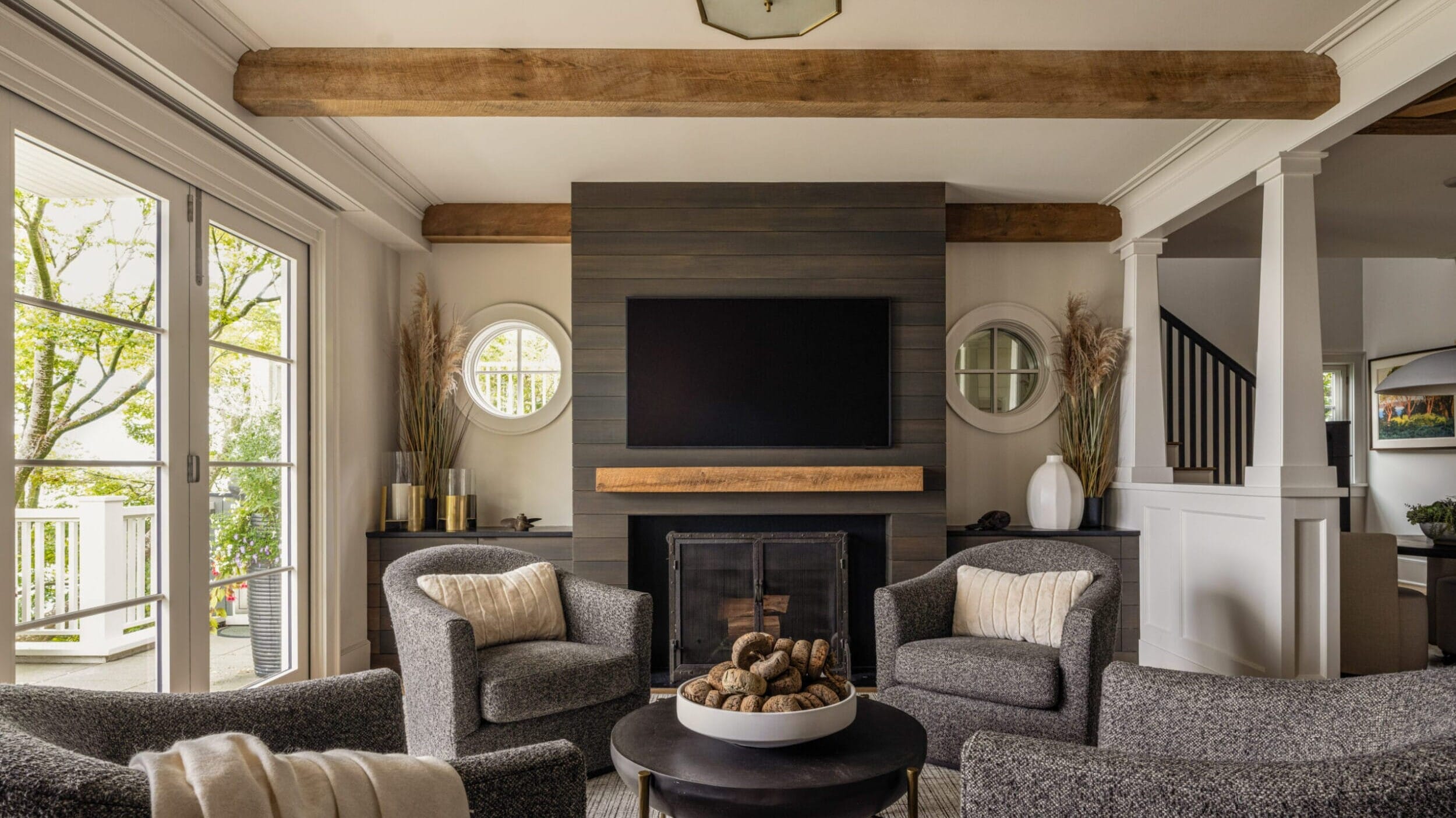 Cozy living room with a fireplace, wall-mounted TV, and four gray armchairs arranged around a round coffee table. Large windows and wooden beams add warmth to the space.