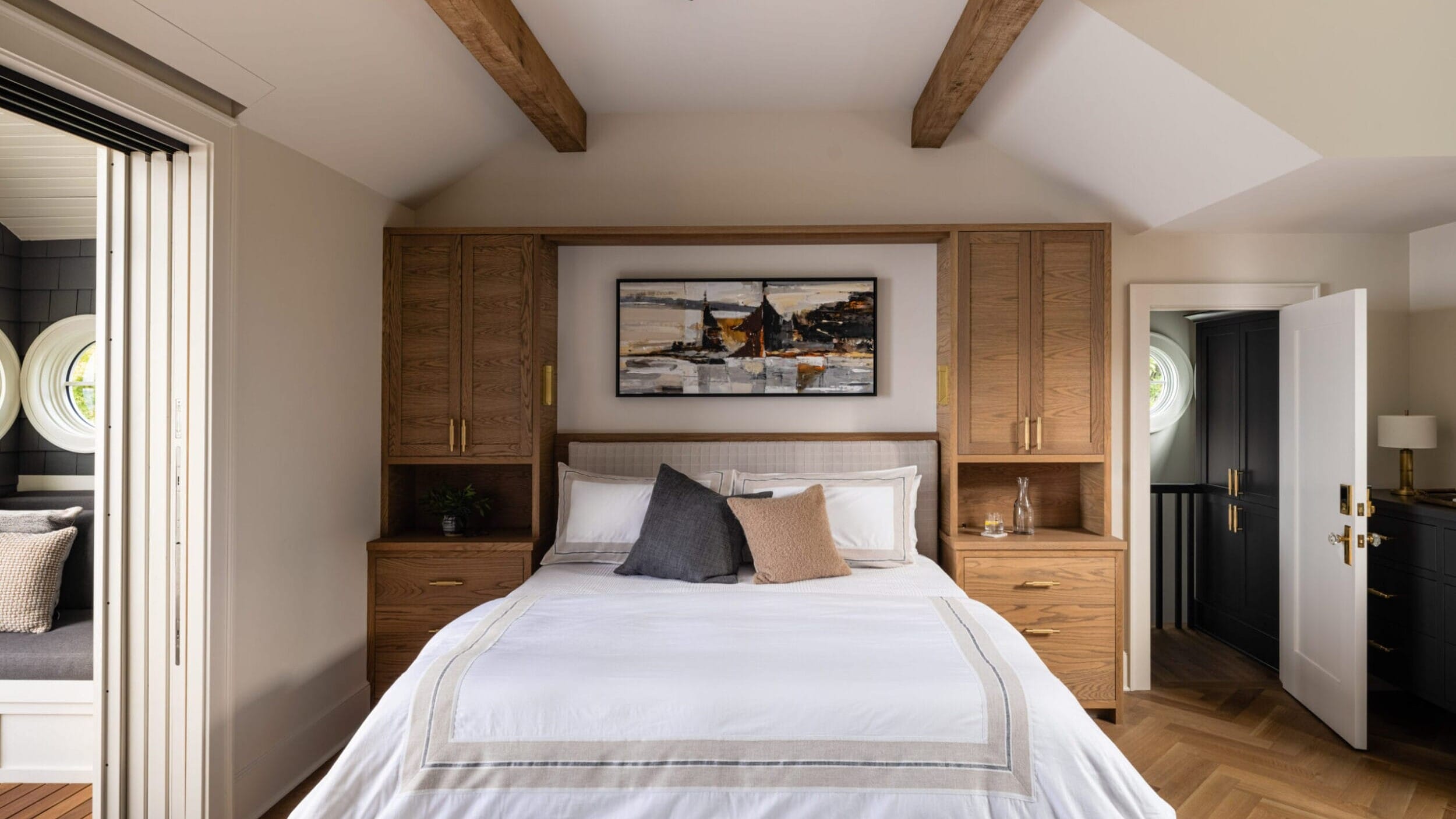 Bedroom with a bed centered against a wooden storage wall, white and gray bedding, decorative pillows, a ceiling fan, exposed beams, and a landscape painting above the bed.