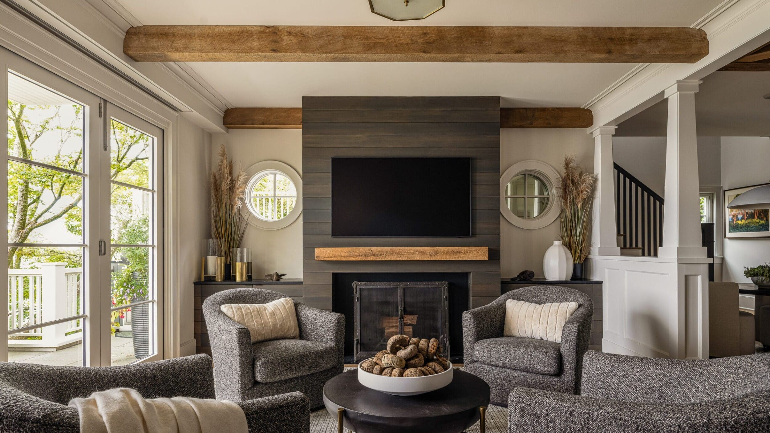 A cozy living room with a fireplace, TV above, round coffee table, and four gray armchairs around it. Exposed wooden beams and large windows create a light, airy atmosphere.