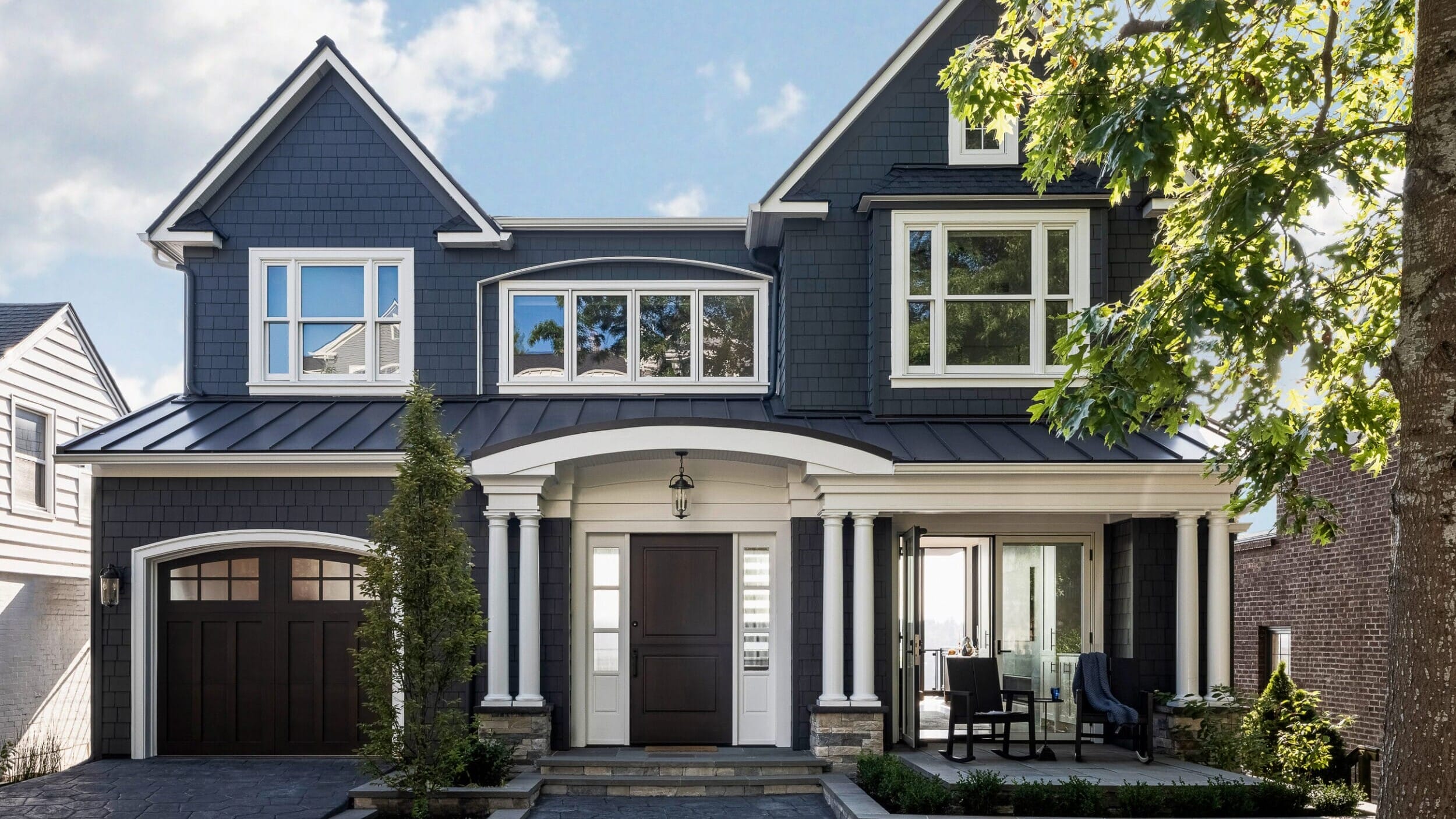 Two-story house with dark gray siding, white trim, and a covered front porch. Double front doors and two-car garage on the left. A tree partially frames the view on the right.