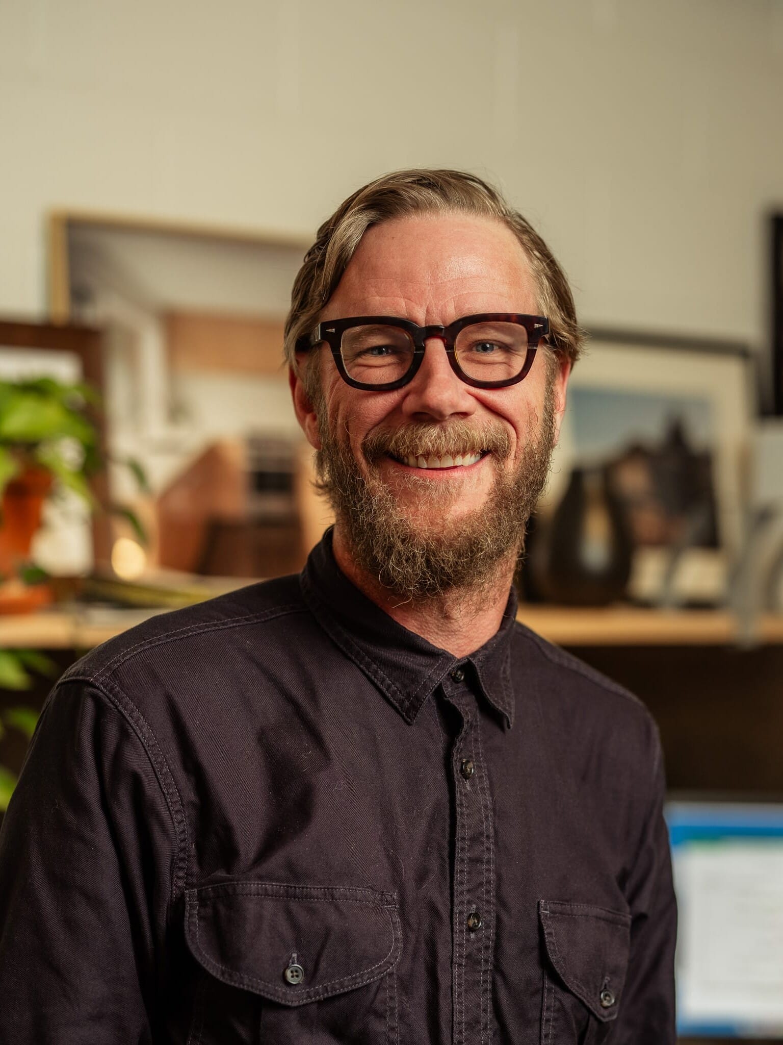 A man with glasses and a beard smiling in front of a desk.